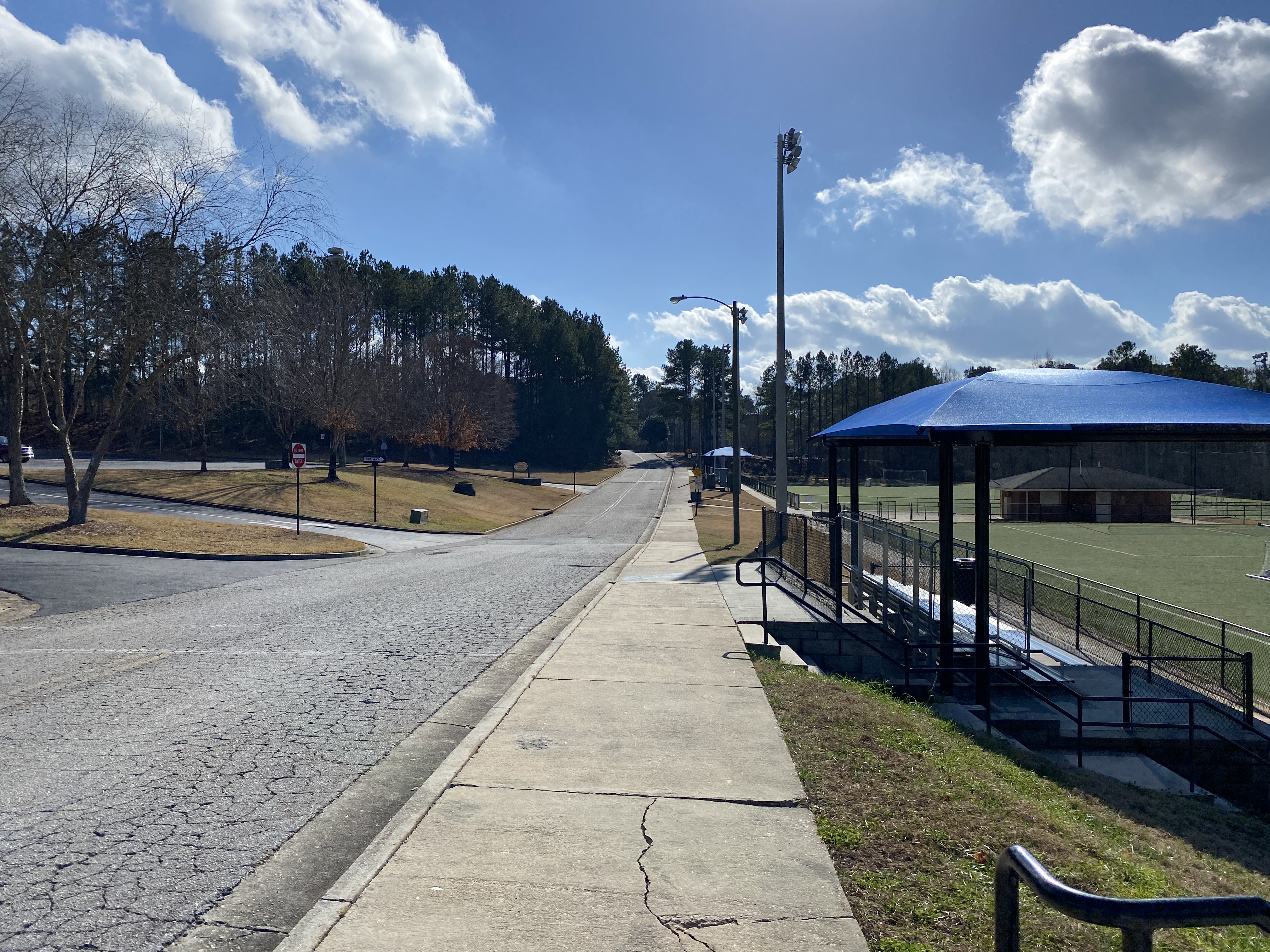 Paved park road with covered bleacher structure, light poles, and trees on a clear day.
