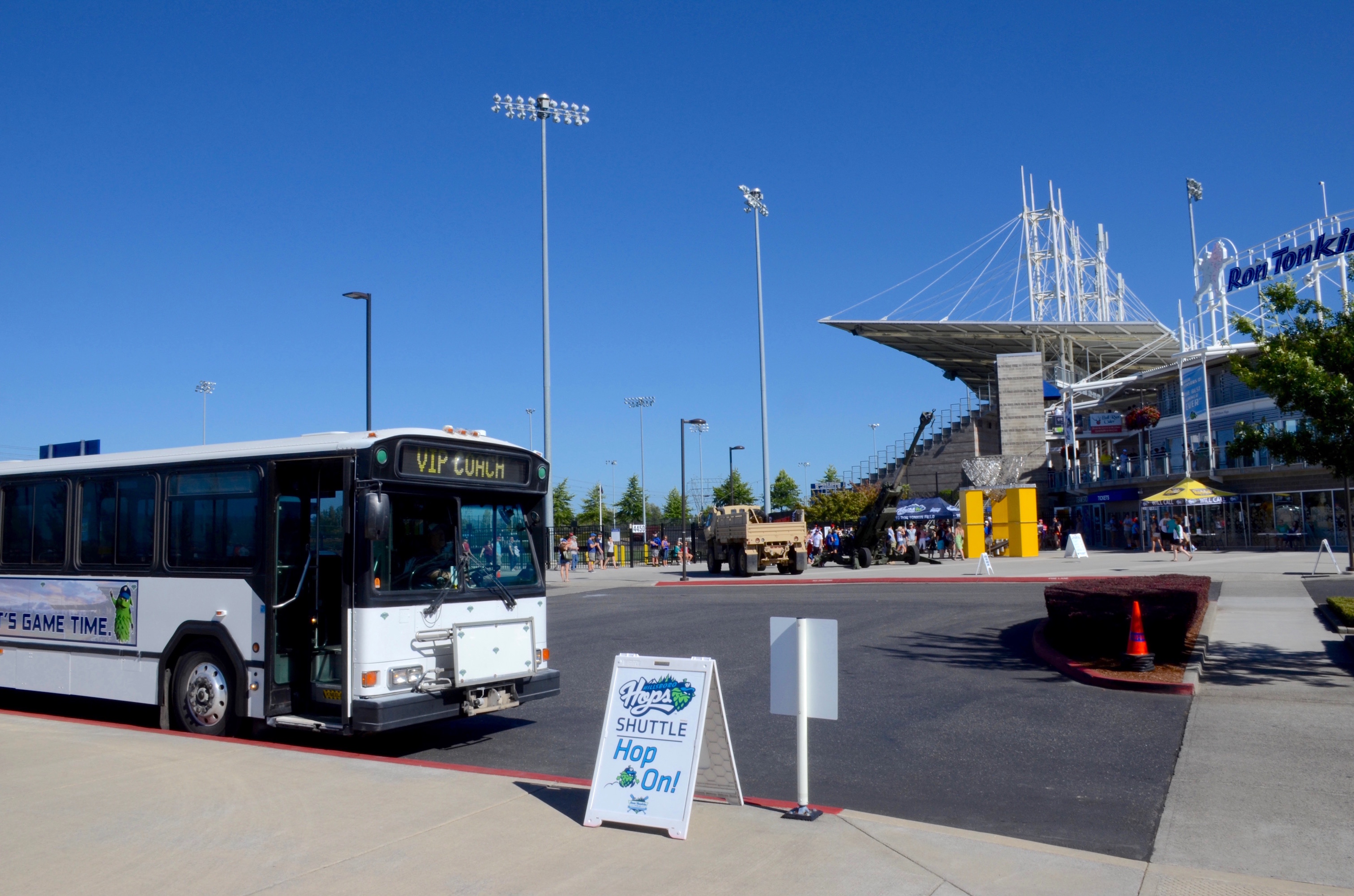 A transit bus marked as a Hops Shuttle parked outside a baseball stadium with light poles visible.