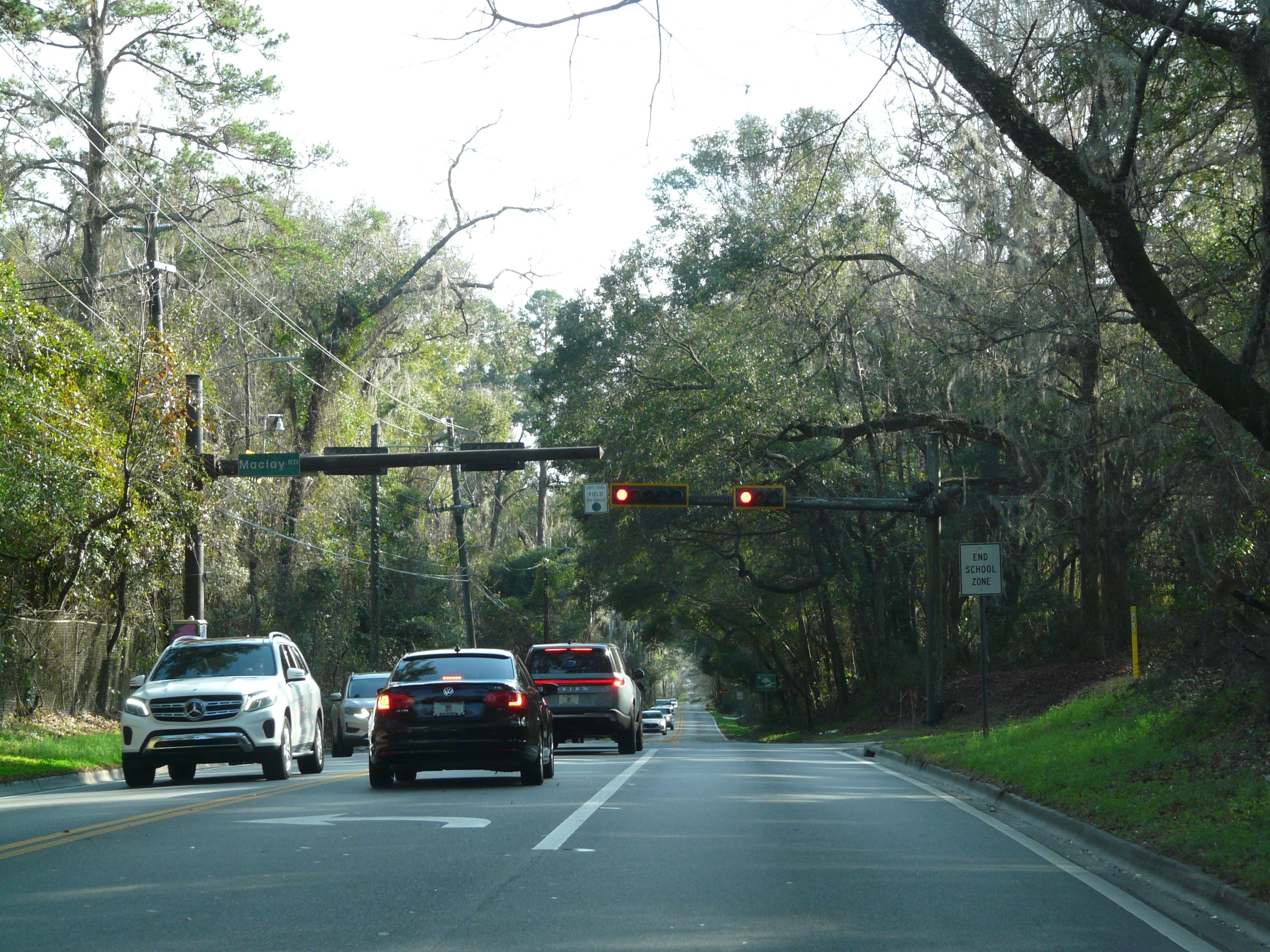Overhead traffic signals showing red lights at a tree-lined road intersection.
