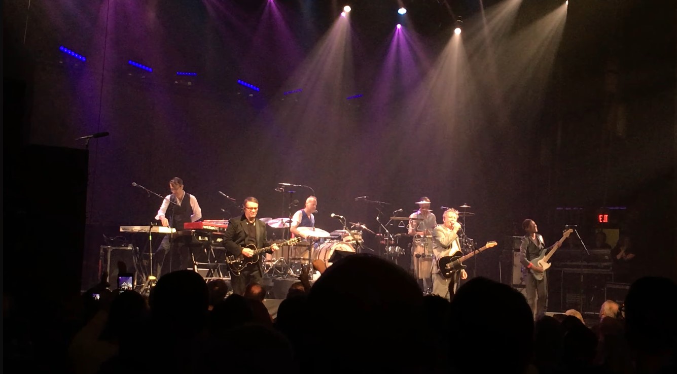 Band on a dimly lit stage with purple lighting and silhouetted audience in the foreground.