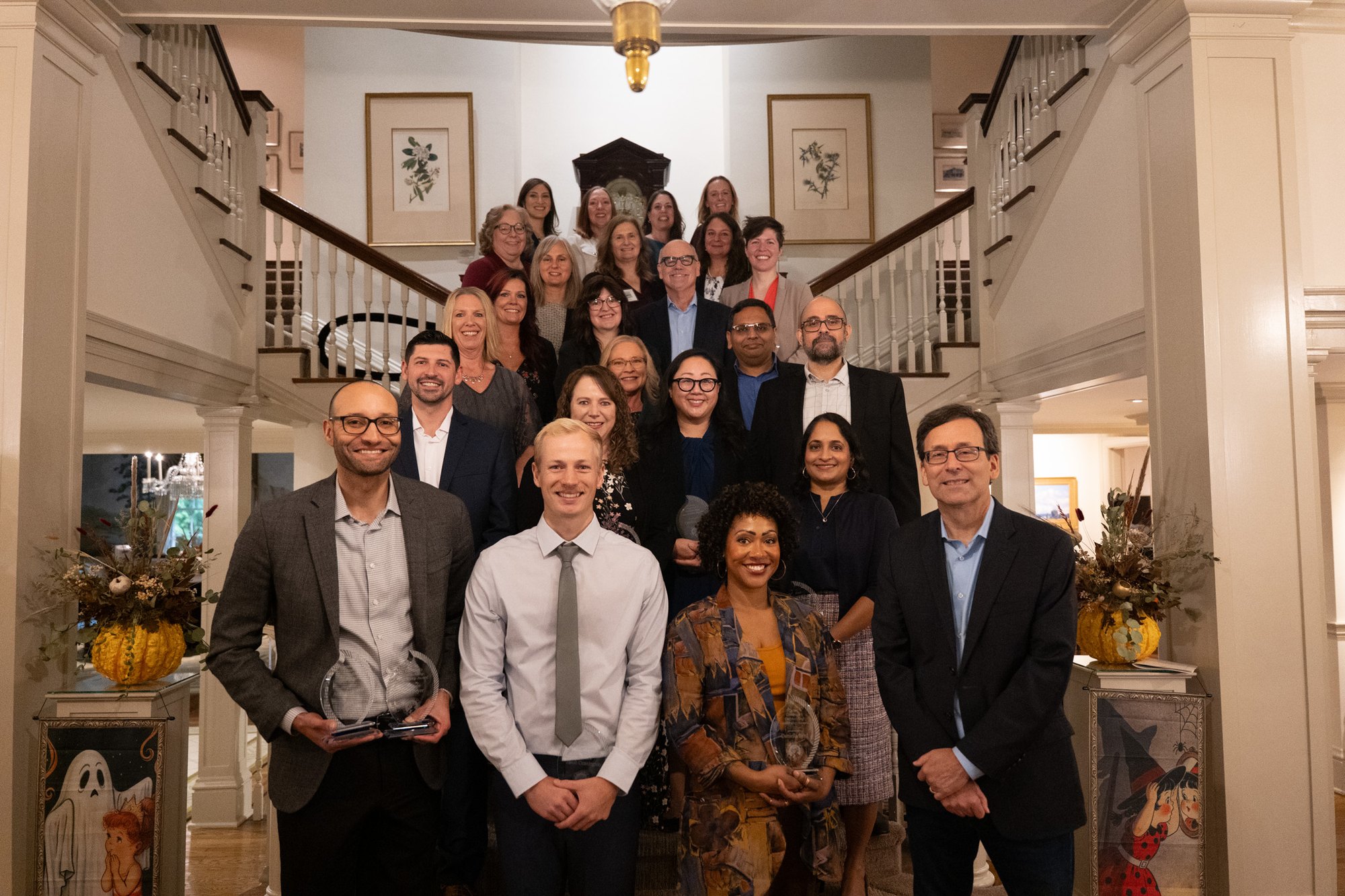 A large group of award recipients pose on a grand staircase, several holding glass trophies.