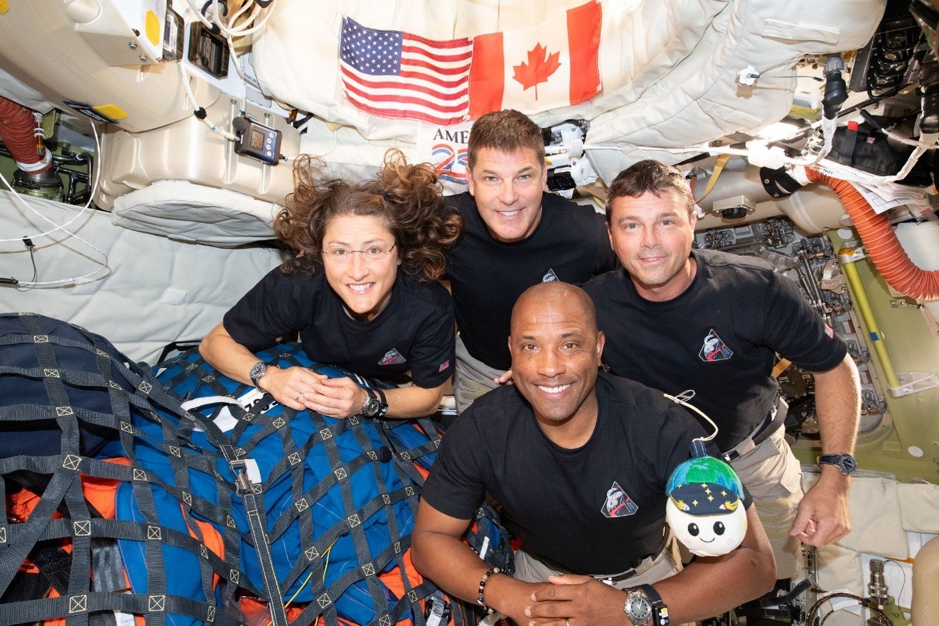 Four astronauts in matching dark shirts float together inside a spacecraft cabin, smiling at the camera.