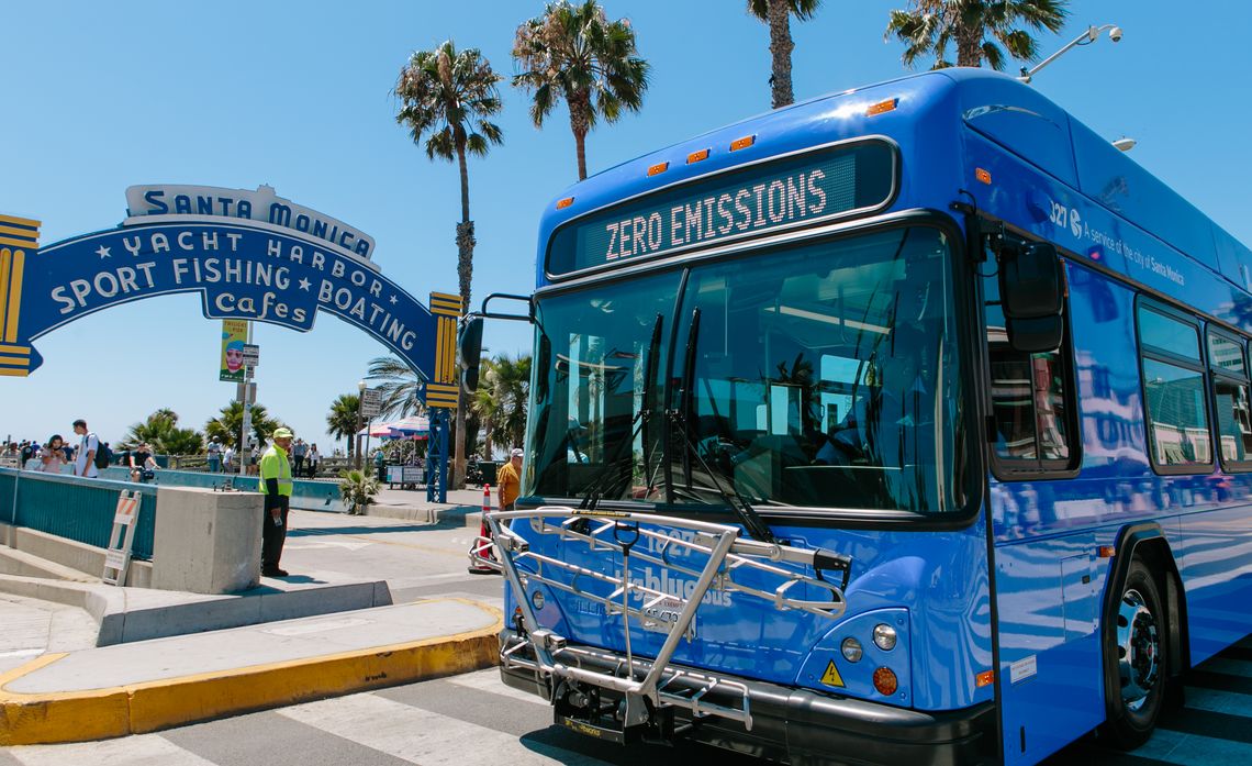 Blue transit bus displaying 'Zero Emissions' parked near a waterfront arch sign with palm trees.