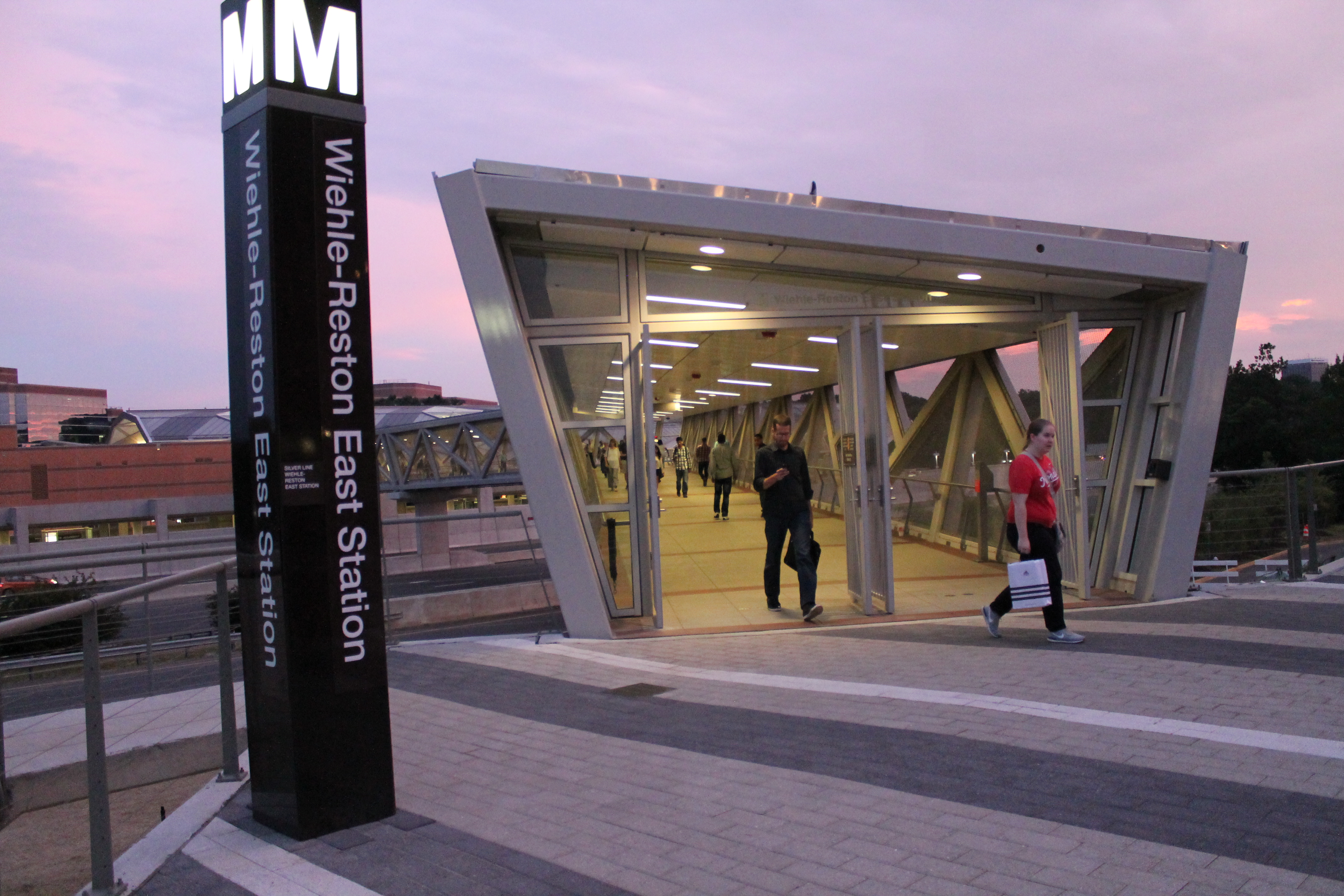Glass-and-steel Metro station entrance at dusk with a tall WMATA sign and pedestrians.
