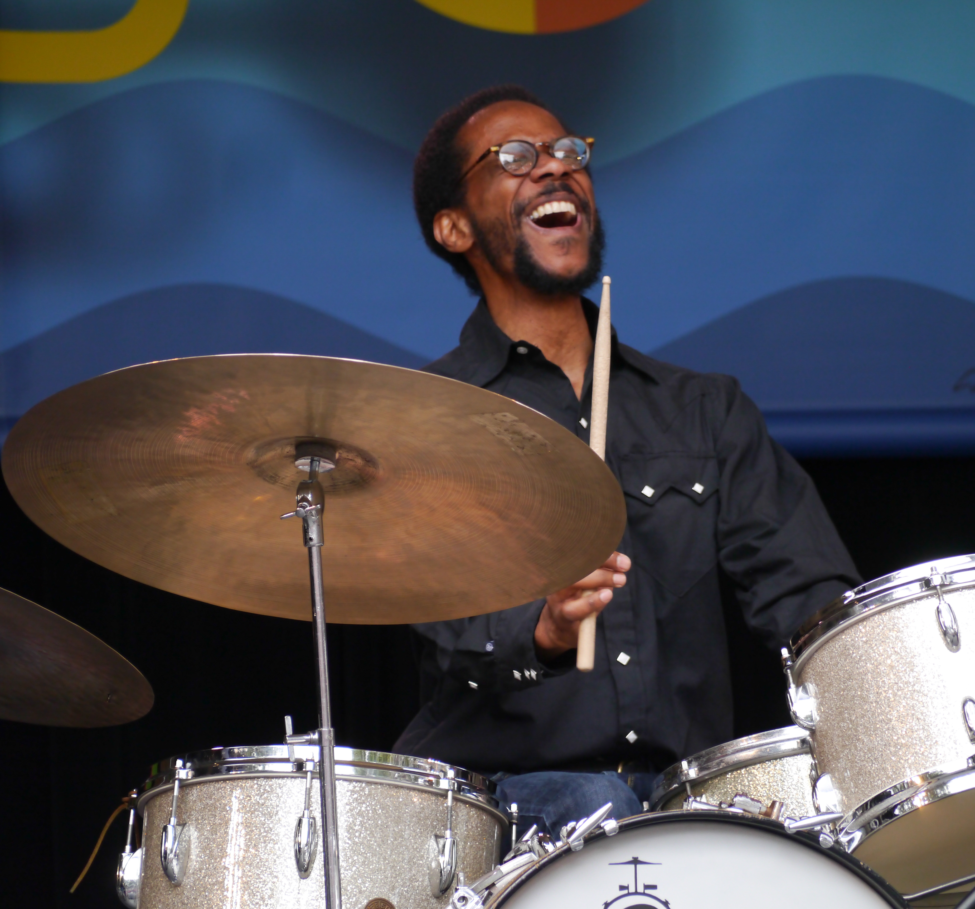 A smiling drummer plays a drum kit on a festival stage with a colorful backdrop.
