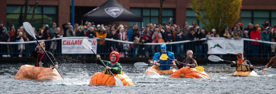 Several costumed paddlers race in giant orange hollowed-out pumpkins on a lake before a large crowd.