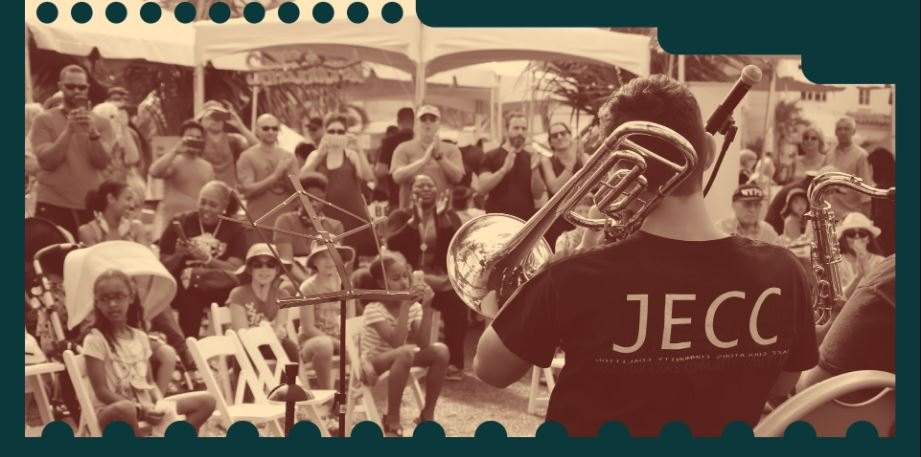 Musician in a JECC t-shirt plays a brass instrument before a large outdoor crowd.