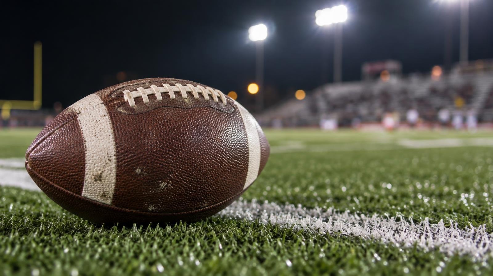 A football resting on green artificial turf near a white yard line under bright stadium lights at night.