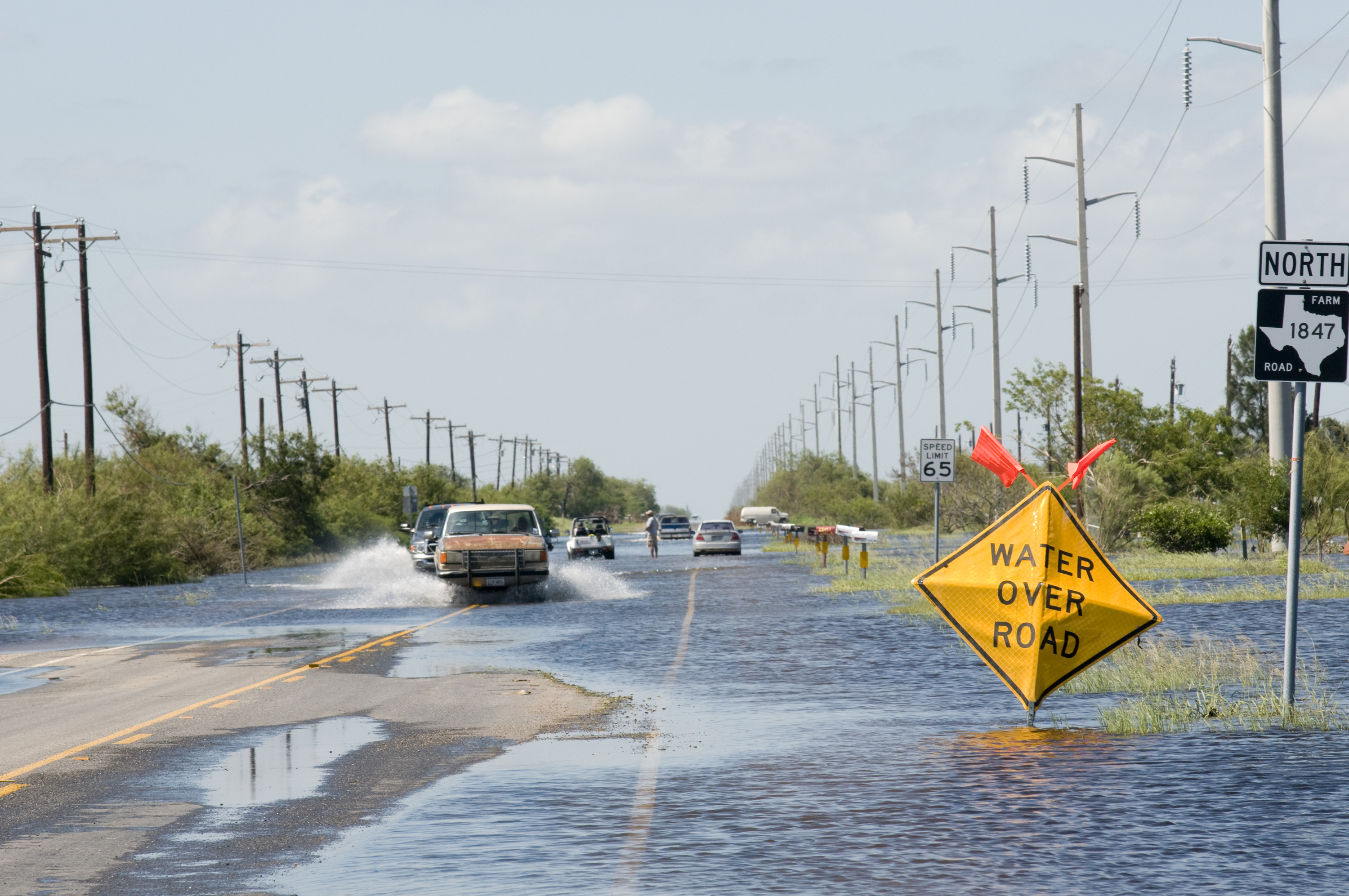 Flooded two-lane road with a yellow water-over-road warning sign and cars driving through standing water.