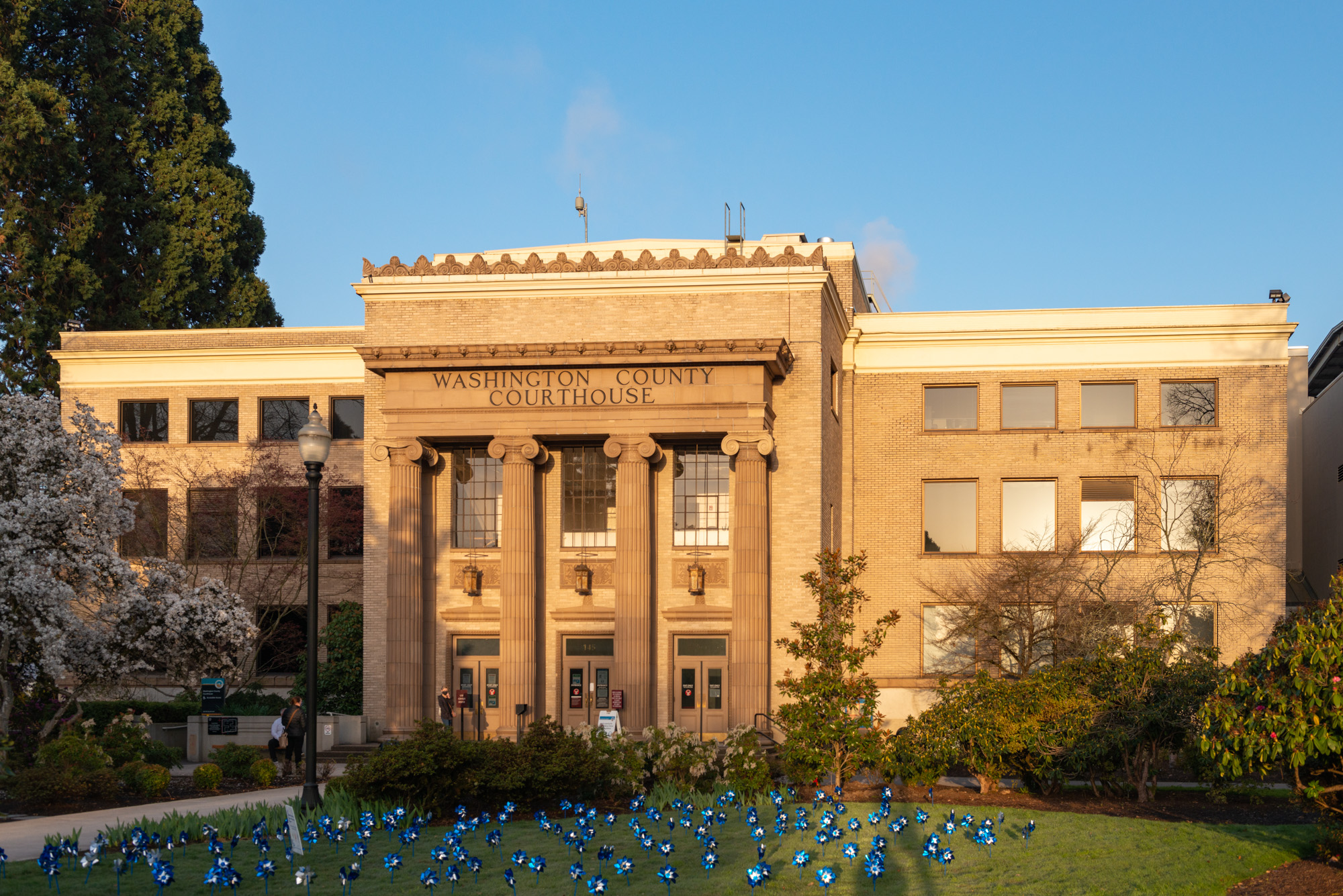 Neoclassical stone courthouse with columns and flowering trees on a sunny day.