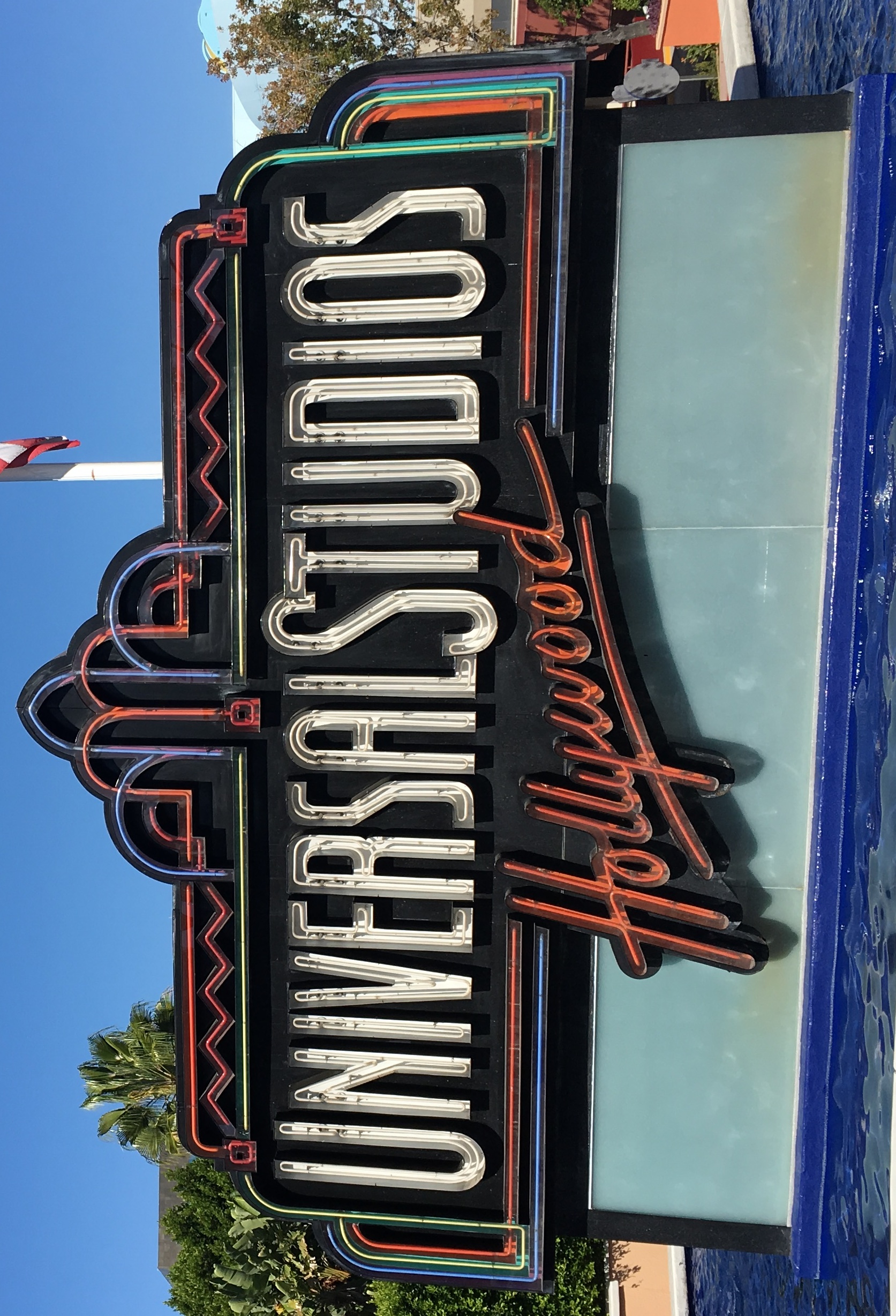 The Universal Studios Hollywood entrance sign flanked by palm trees under a clear blue sky.