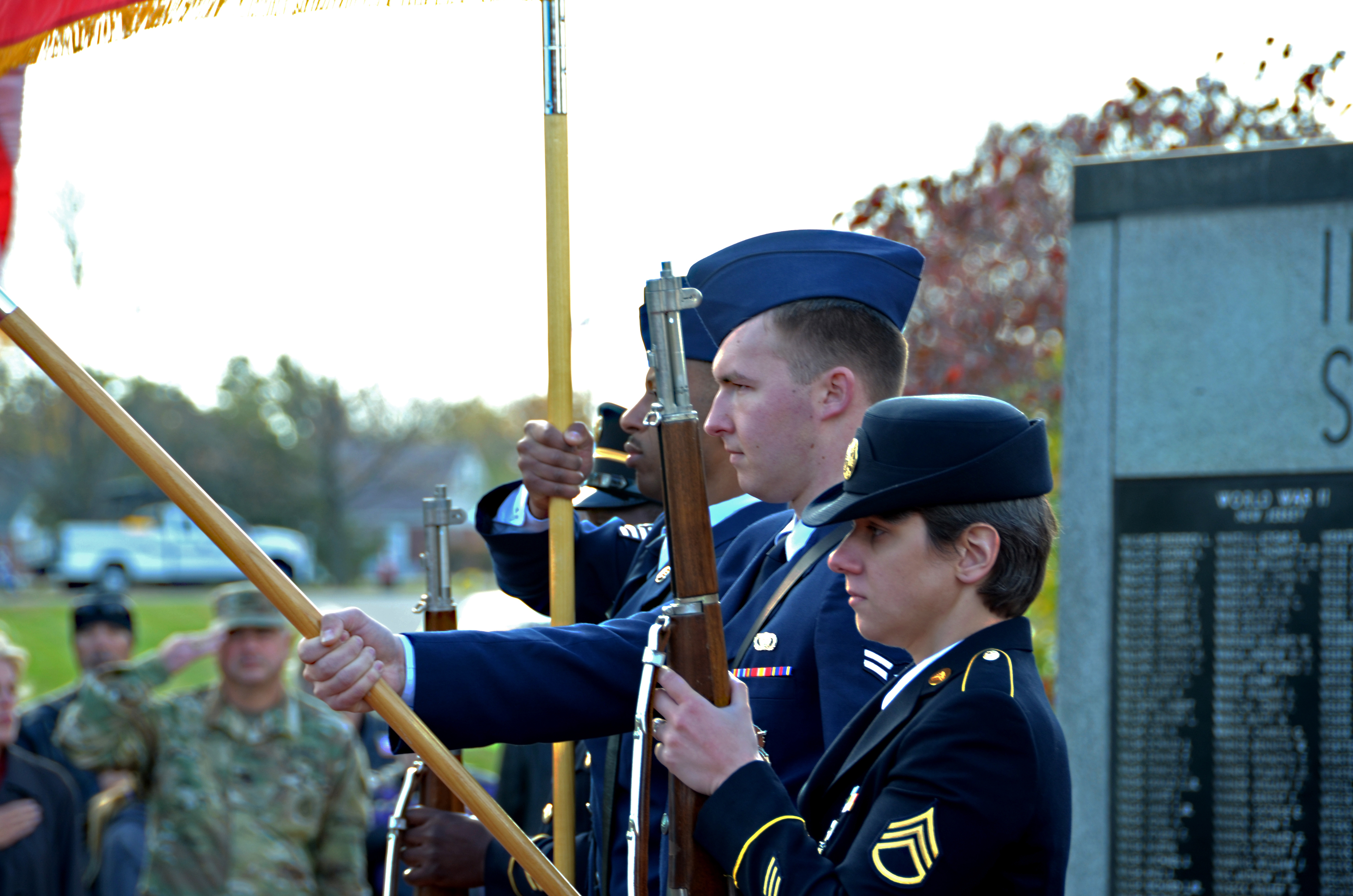 Military color guard in dress uniforms presenting flags at an outdoor Veterans Day ceremony.