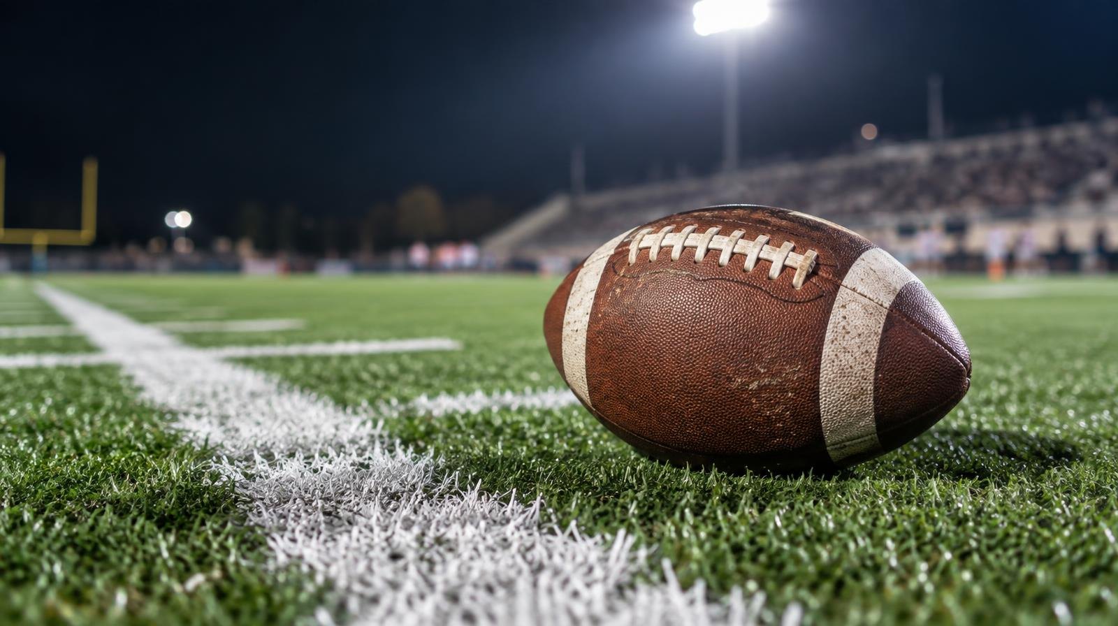 An American football resting on green turf beside a white painted yard line.