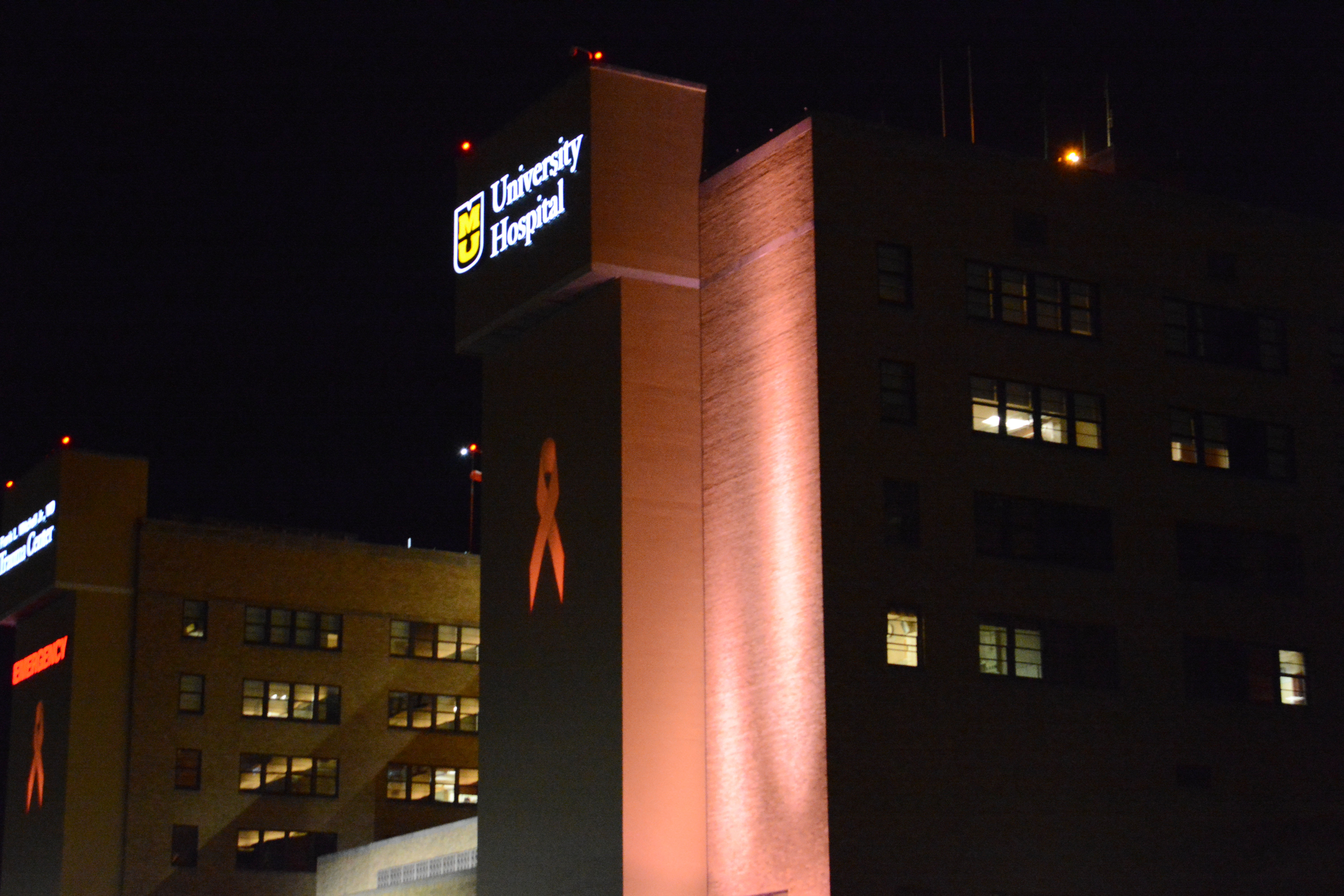 Hospital building at night lit in orange with two large pink awareness ribbons on its exterior walls.