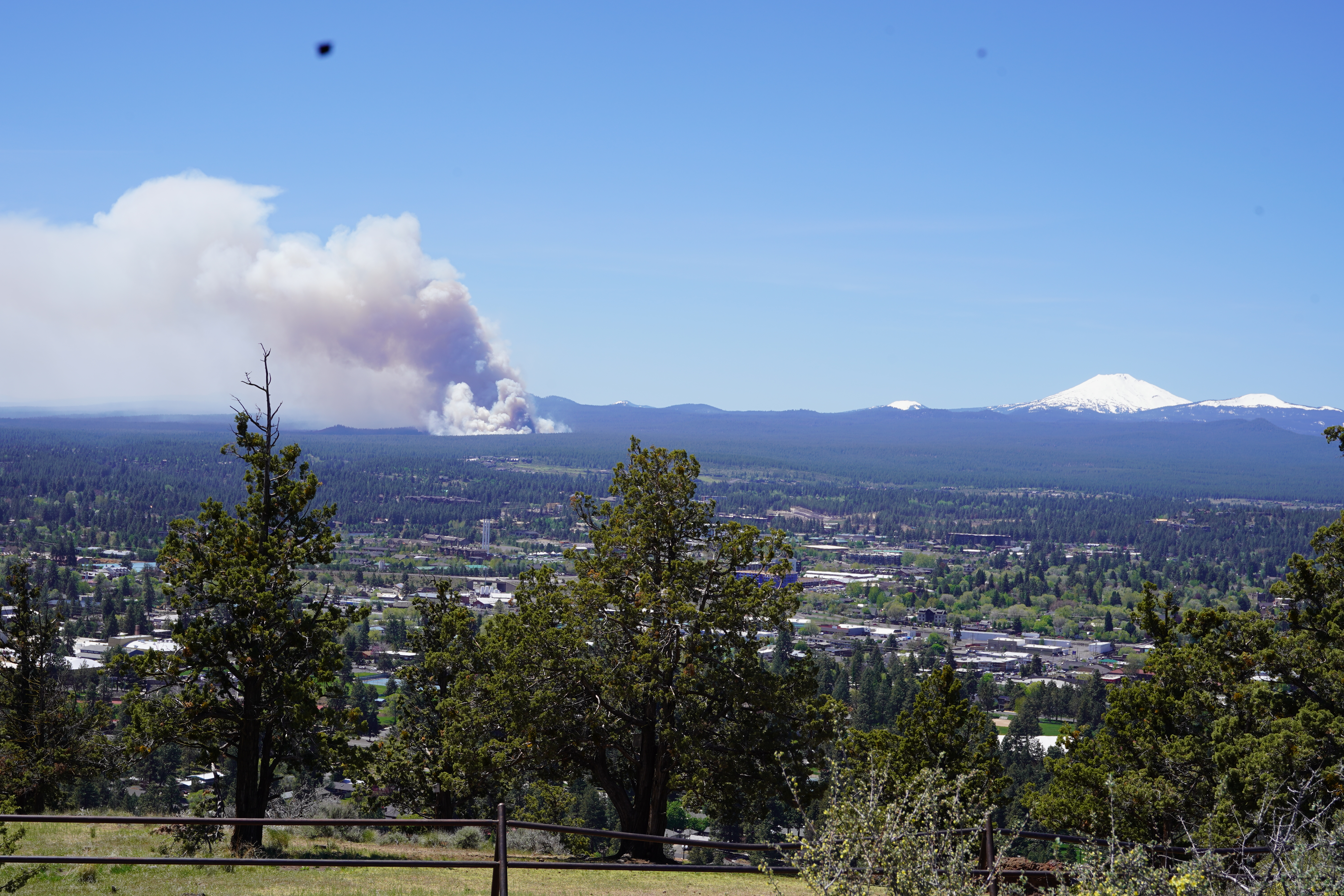 Massive smoke plume rising over forested hills near a town, mountains in background.