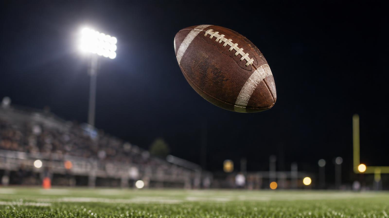 A football spiraling in mid-air above a grass field under bright stadium lights at night.