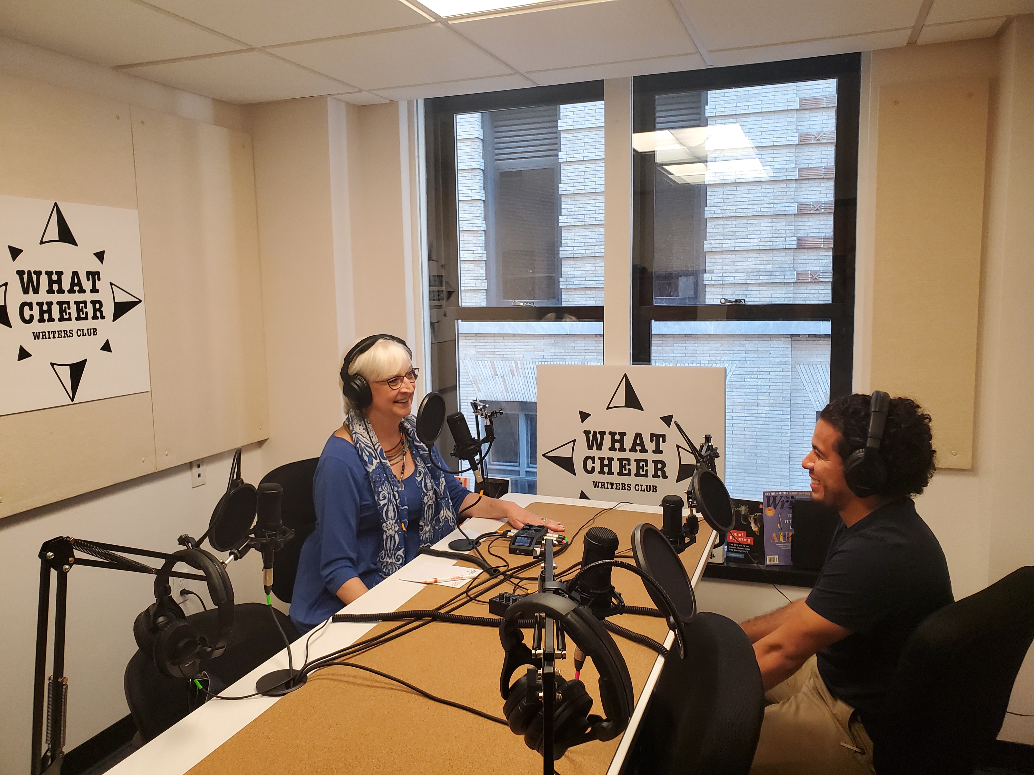 Two people face each other across a desk with microphones and headphones in a podcast studio.