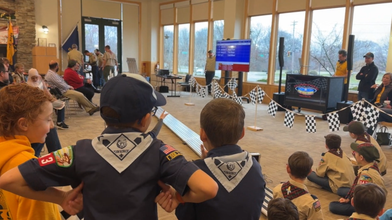 Children in Cub Scout uniforms seated in a large hall watching a presentation screen.
