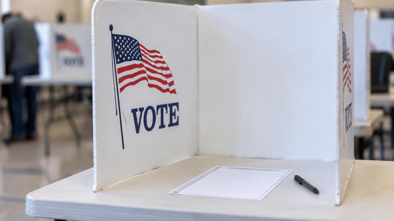 A white voting booth on a table with a blank ballot and pen inside a polling place.