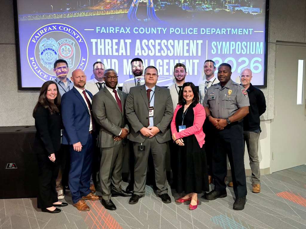 Group of people posing in front of a Threat Assessment and Management Symposium banner.