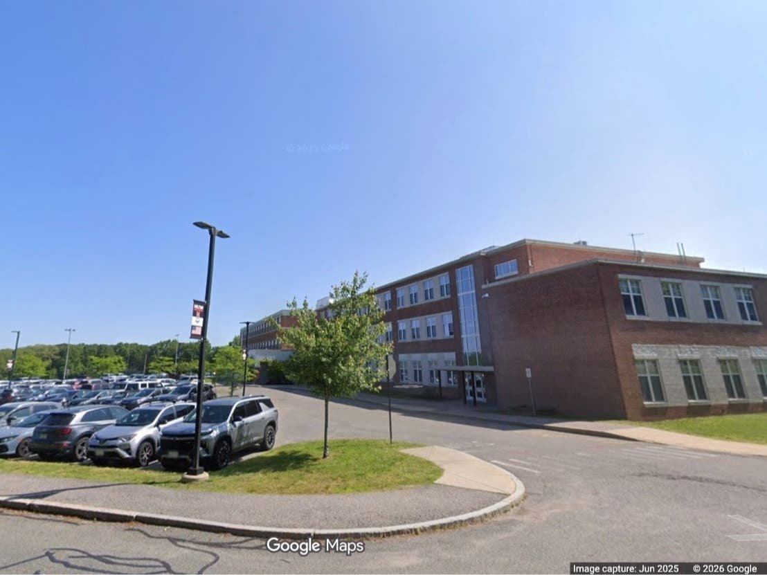 Multi-story brick school building with a parking lot in the foreground under a clear blue sky.