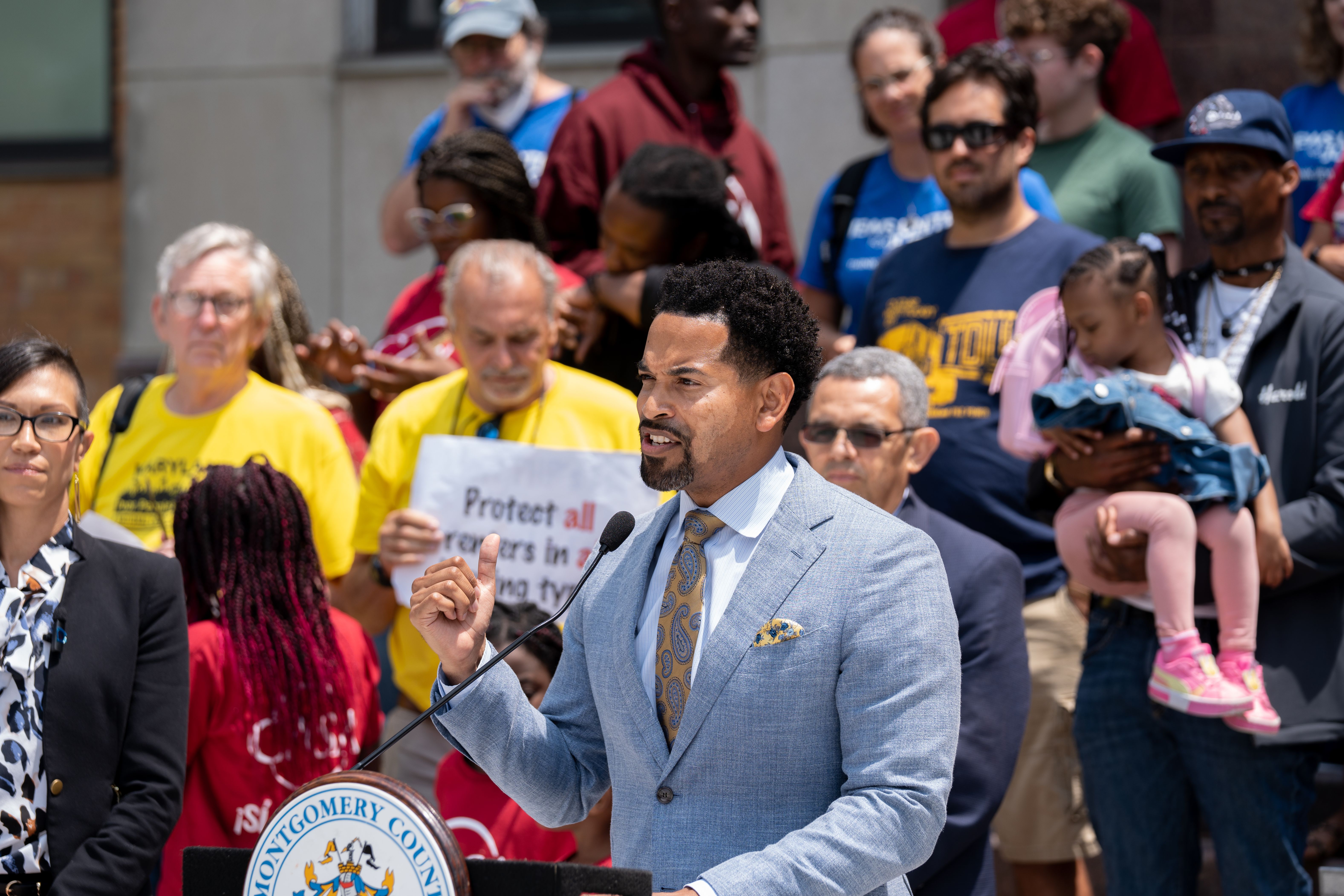 Man in a light blue suit speaks at an outdoor podium before a crowd.