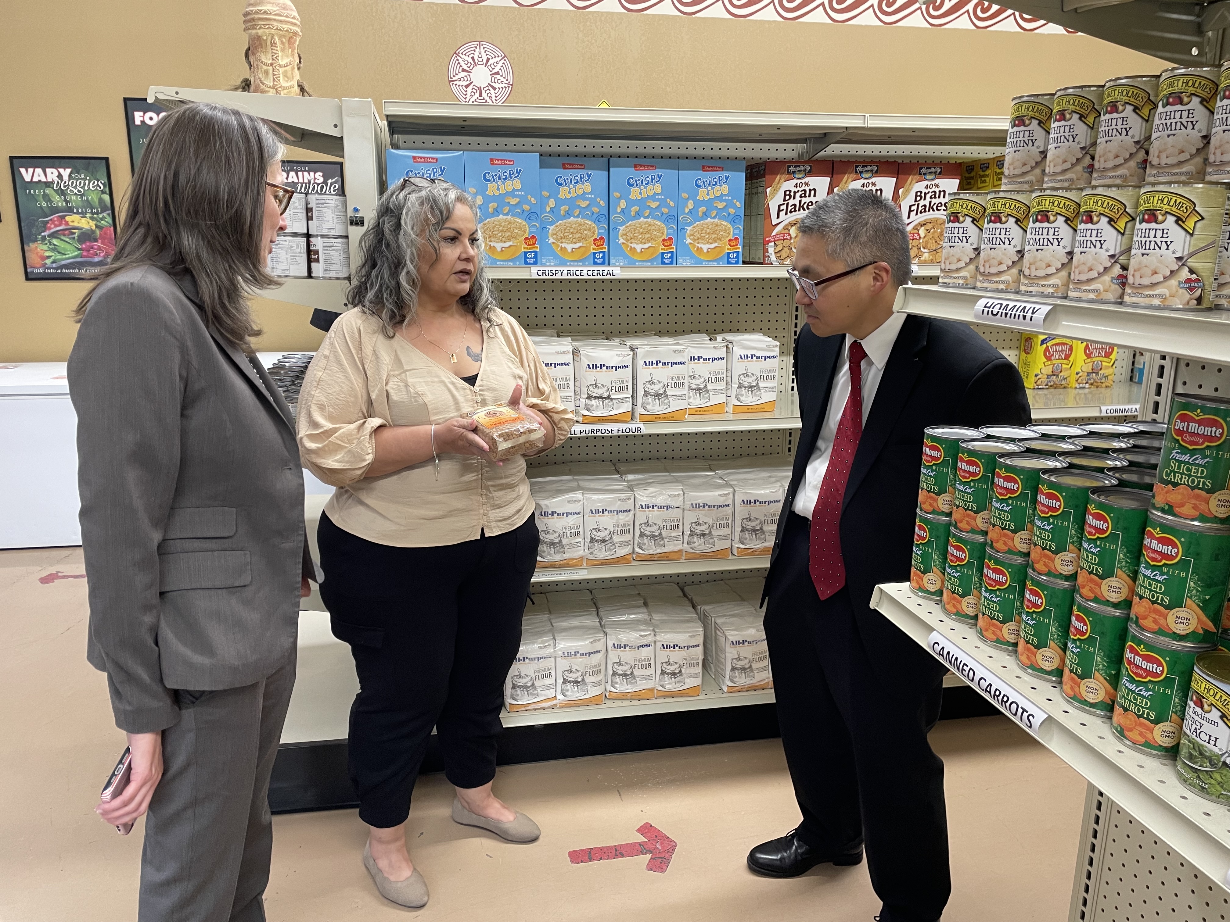 Three people stand before shelves lined with canned goods and packaged food in a food pantry.