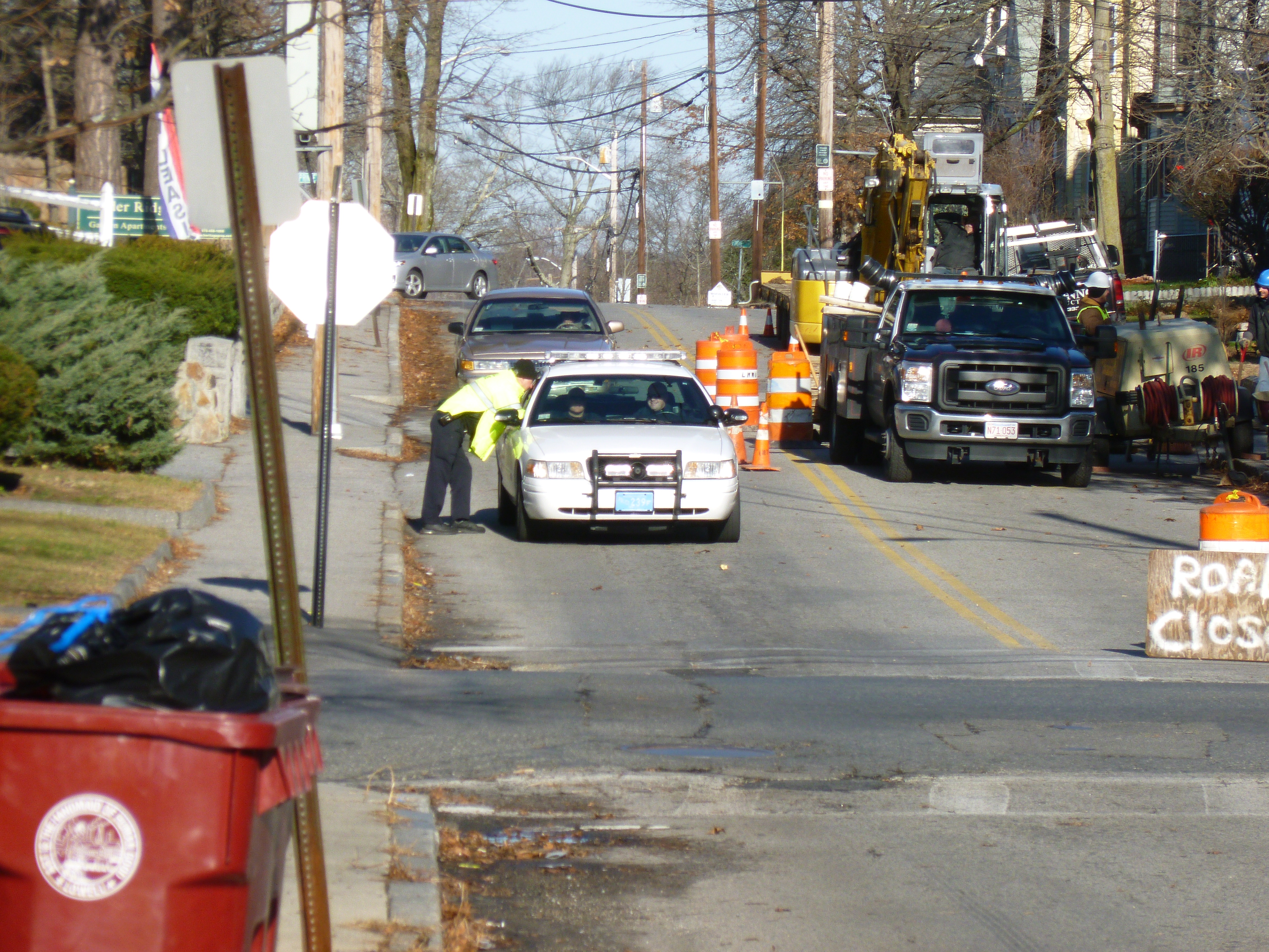 A uniformed police officer in a yellow safety vest stands beside a white patrol car on a road.