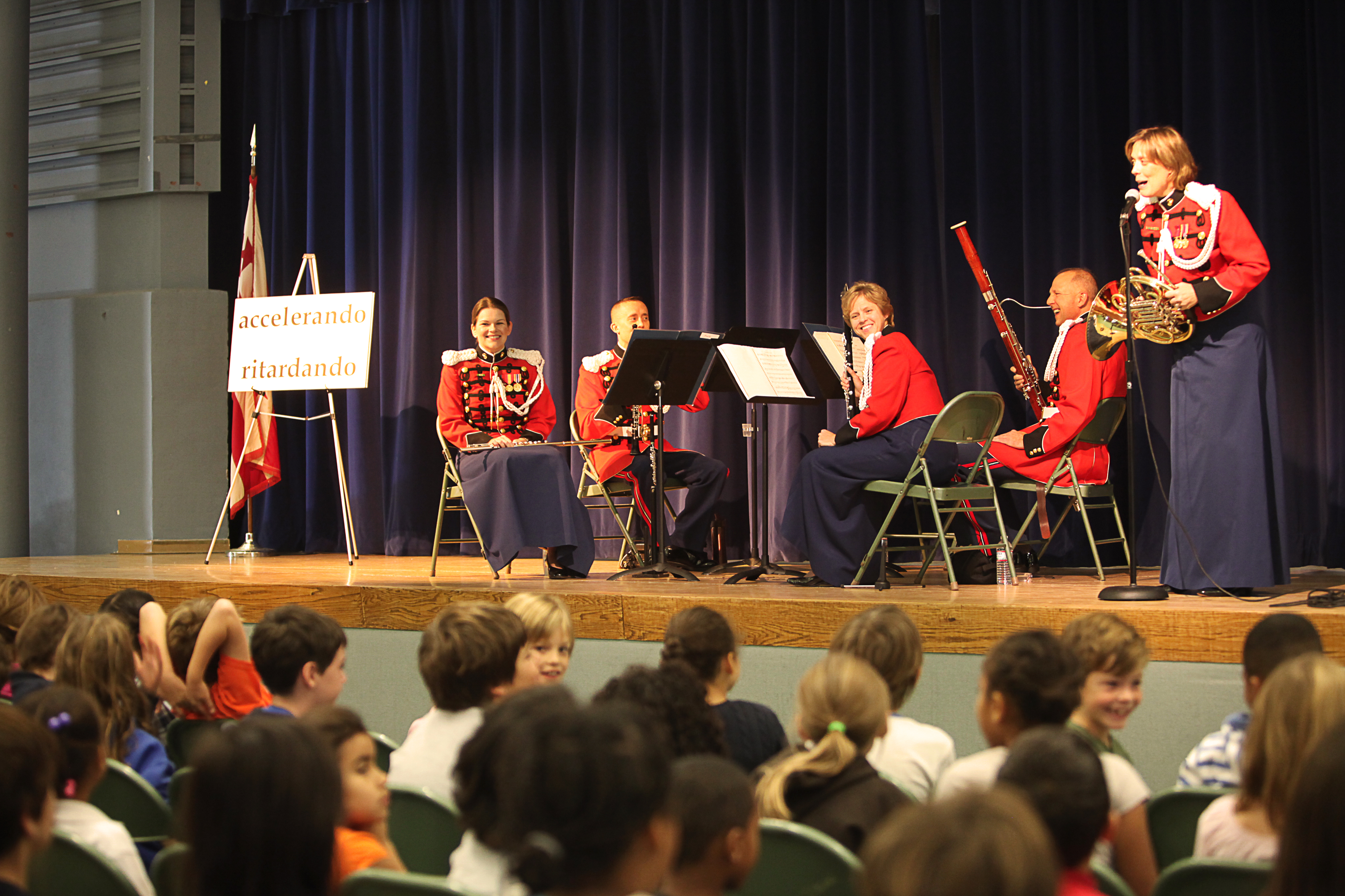 Musicians in red uniforms perform on a stage for a seated audience of children in a school auditorium.