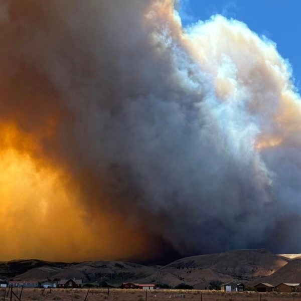 Towering orange and gray smoke columns rise from a wildfire over arid hills.