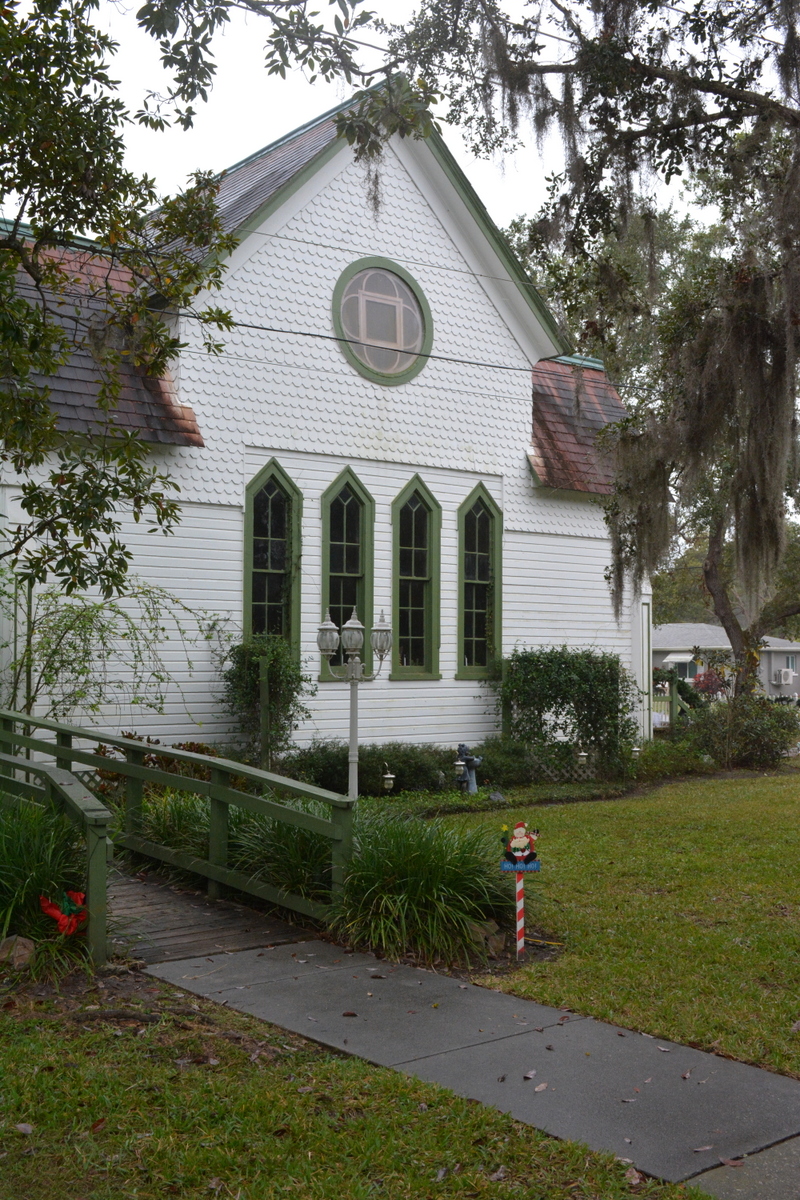 White Carpenter Gothic chapel with green trim and arched windows surrounded by Spanish moss-draped trees.