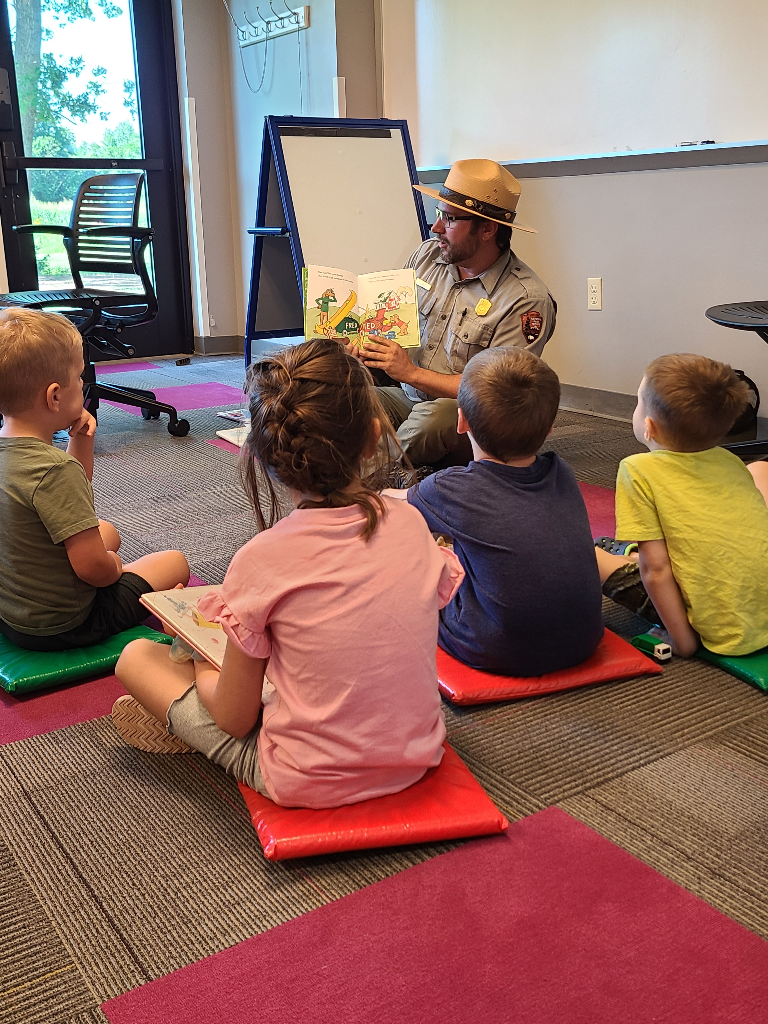Adult in uniform reading a colorful picture book to young children on floor mats indoors.