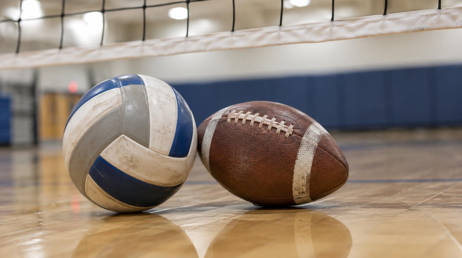 A volleyball and football resting on a hardwood gym floor near a net.