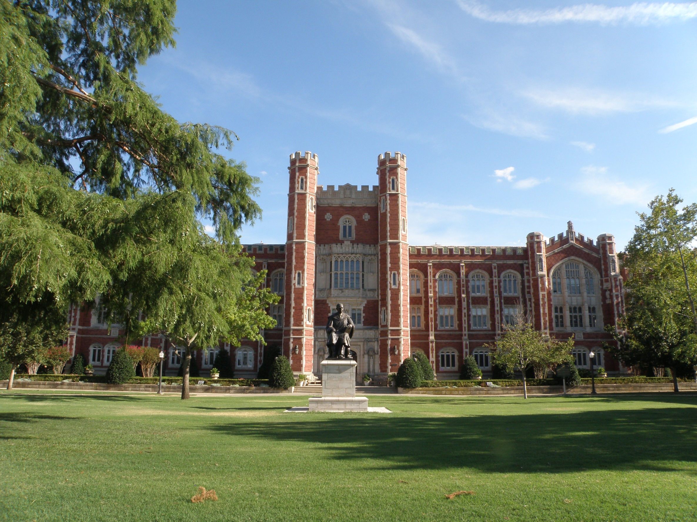 Red-brick Gothic Revival university building with crenellated towers, a bronze statue on the lawn, and mature trees.