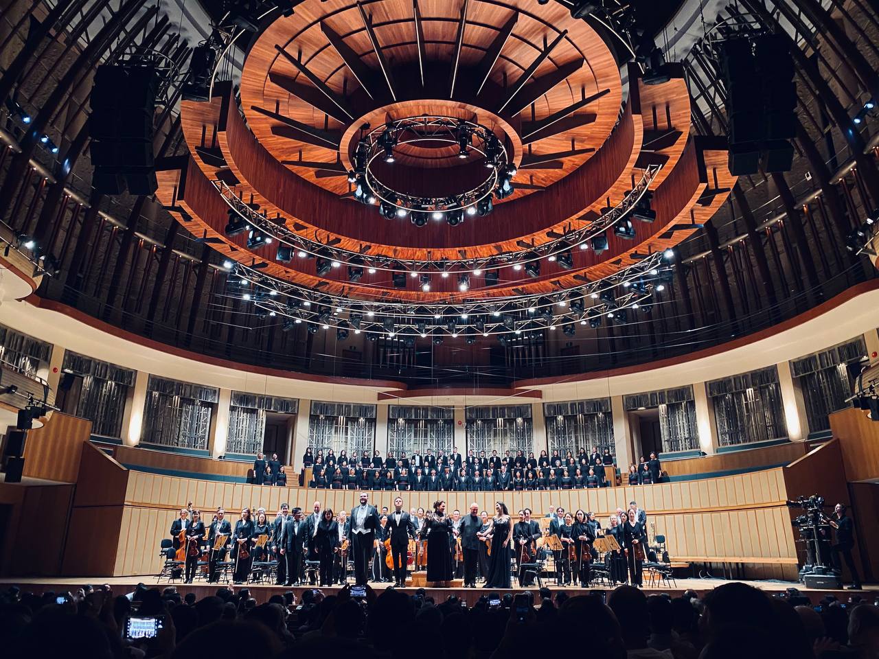 Orchestra and chorus on a lit concert stage bowing before a full audience in a grand hall.