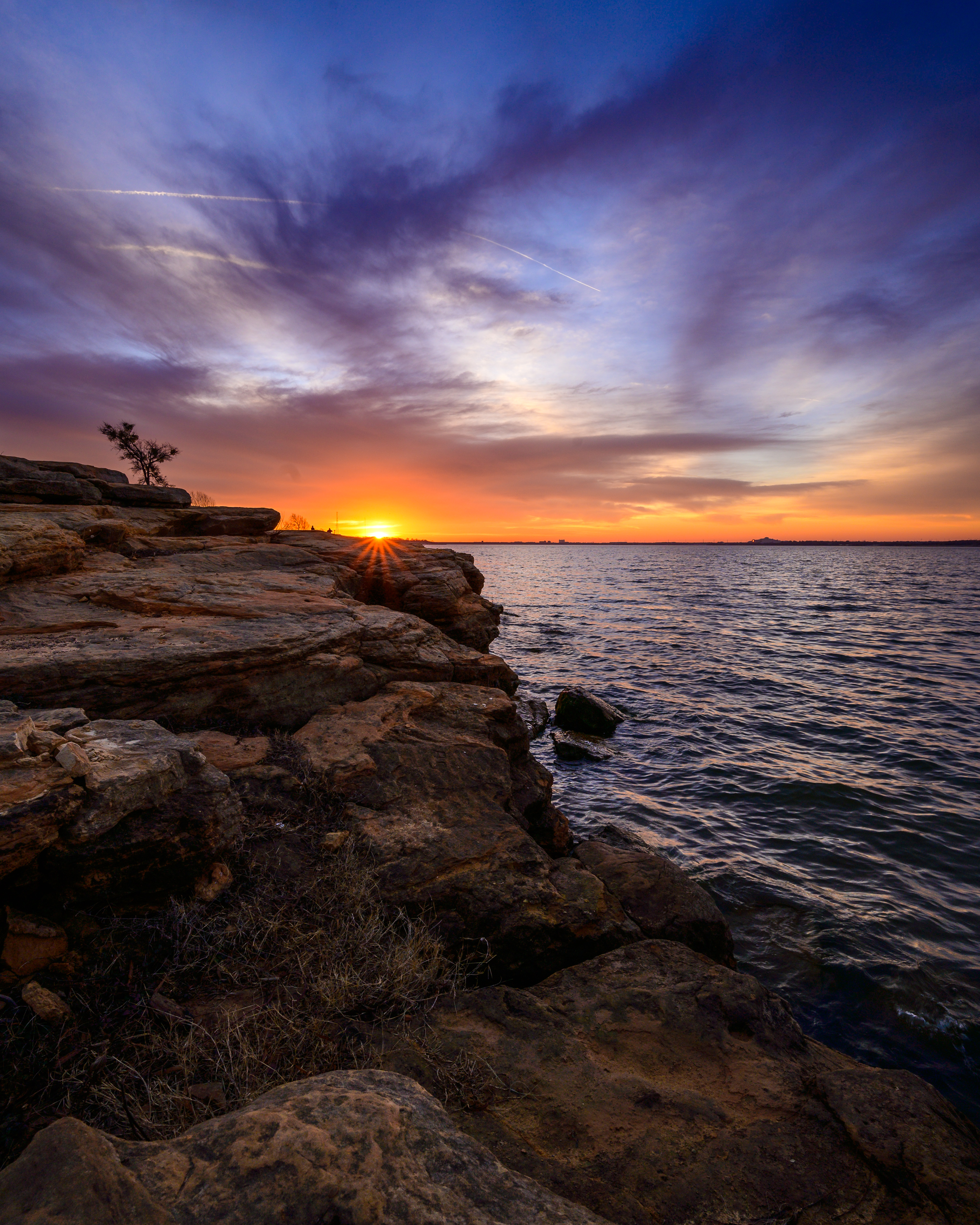 Rocky shoreline at a lake at sunset with vivid orange and purple sky reflected in calm water.