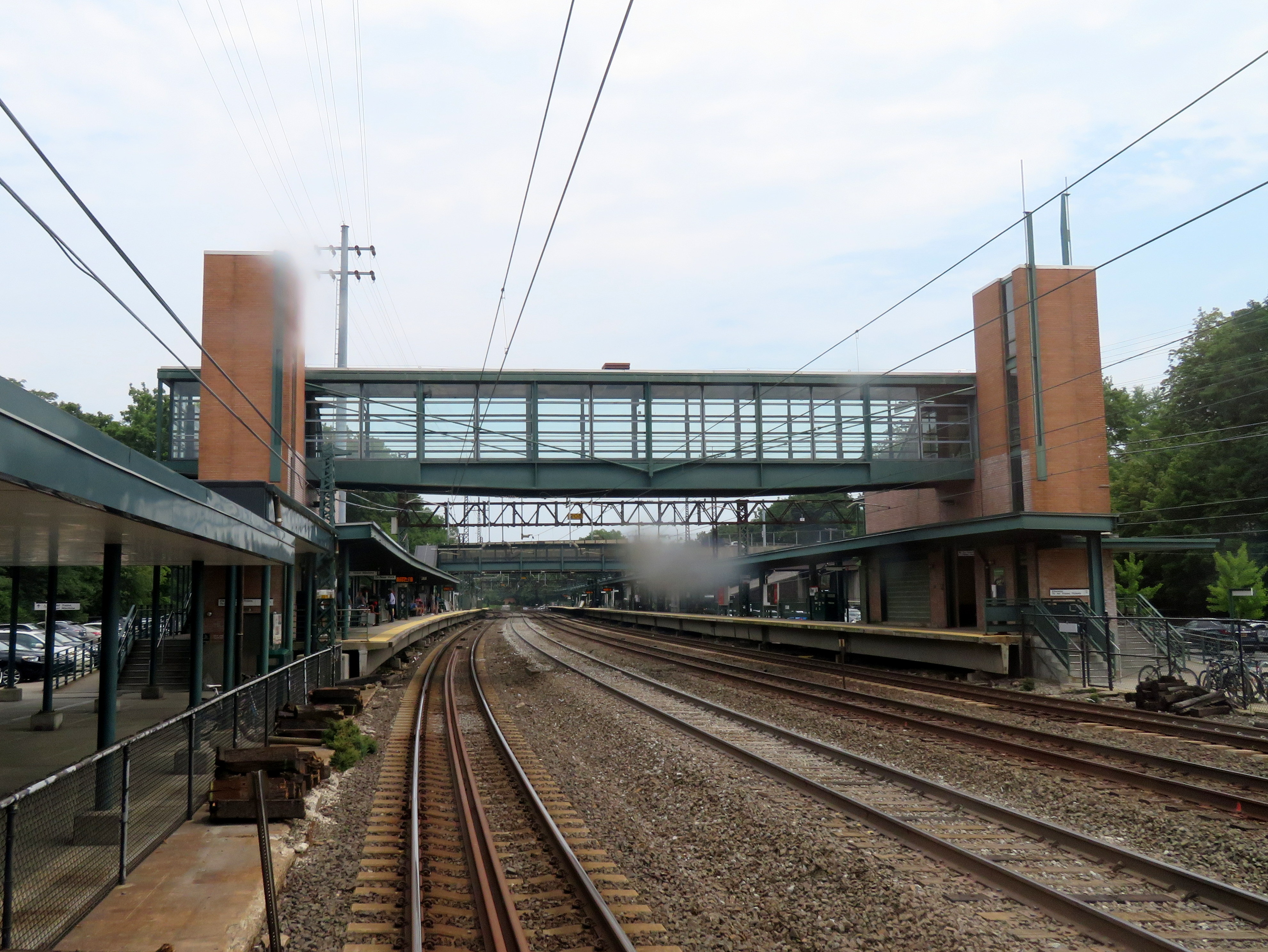 Train station platforms with glass pedestrian overpass and overhead catenary wires.