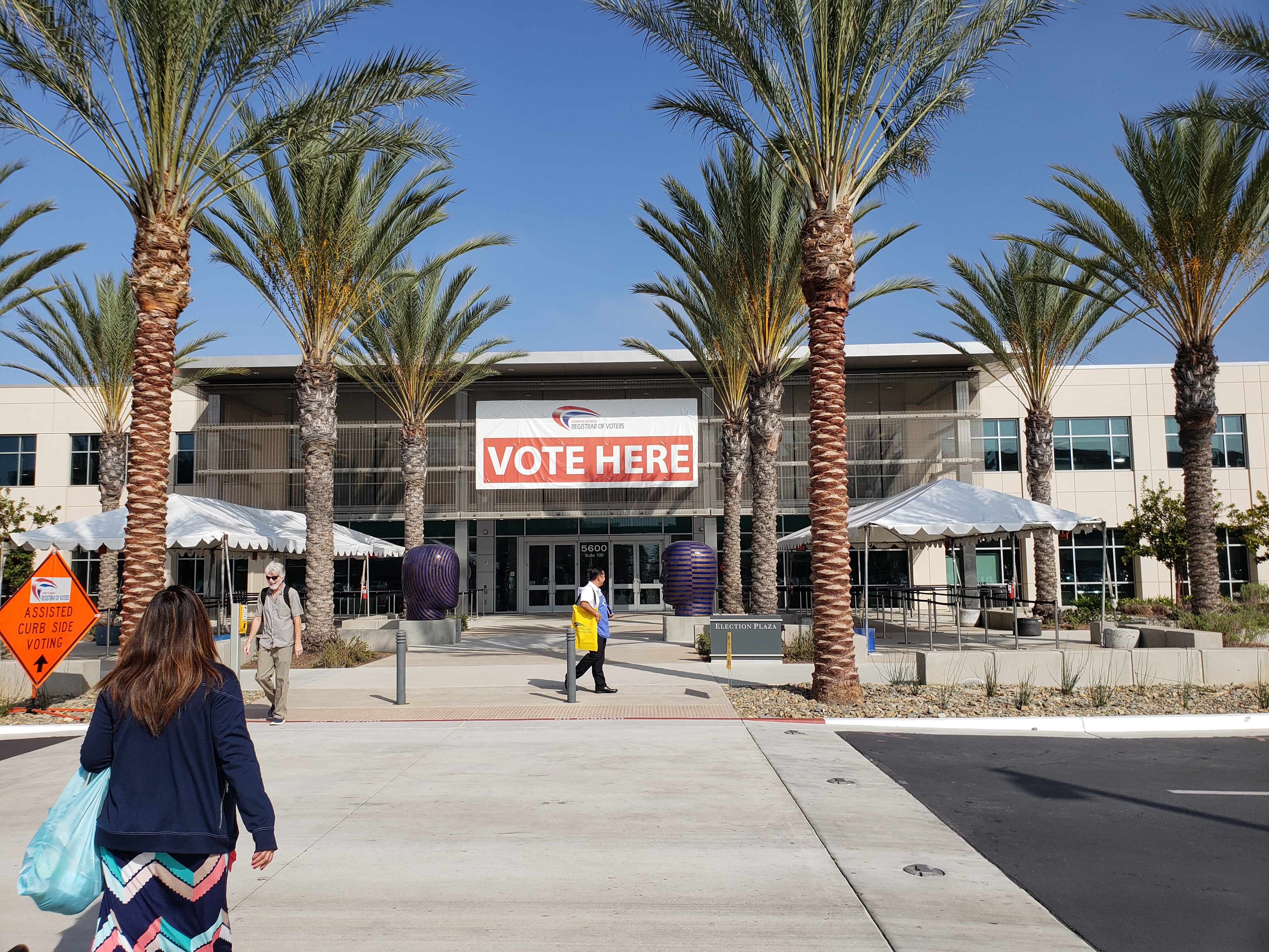 Exterior of a government building with a large Vote Here banner and palm trees.