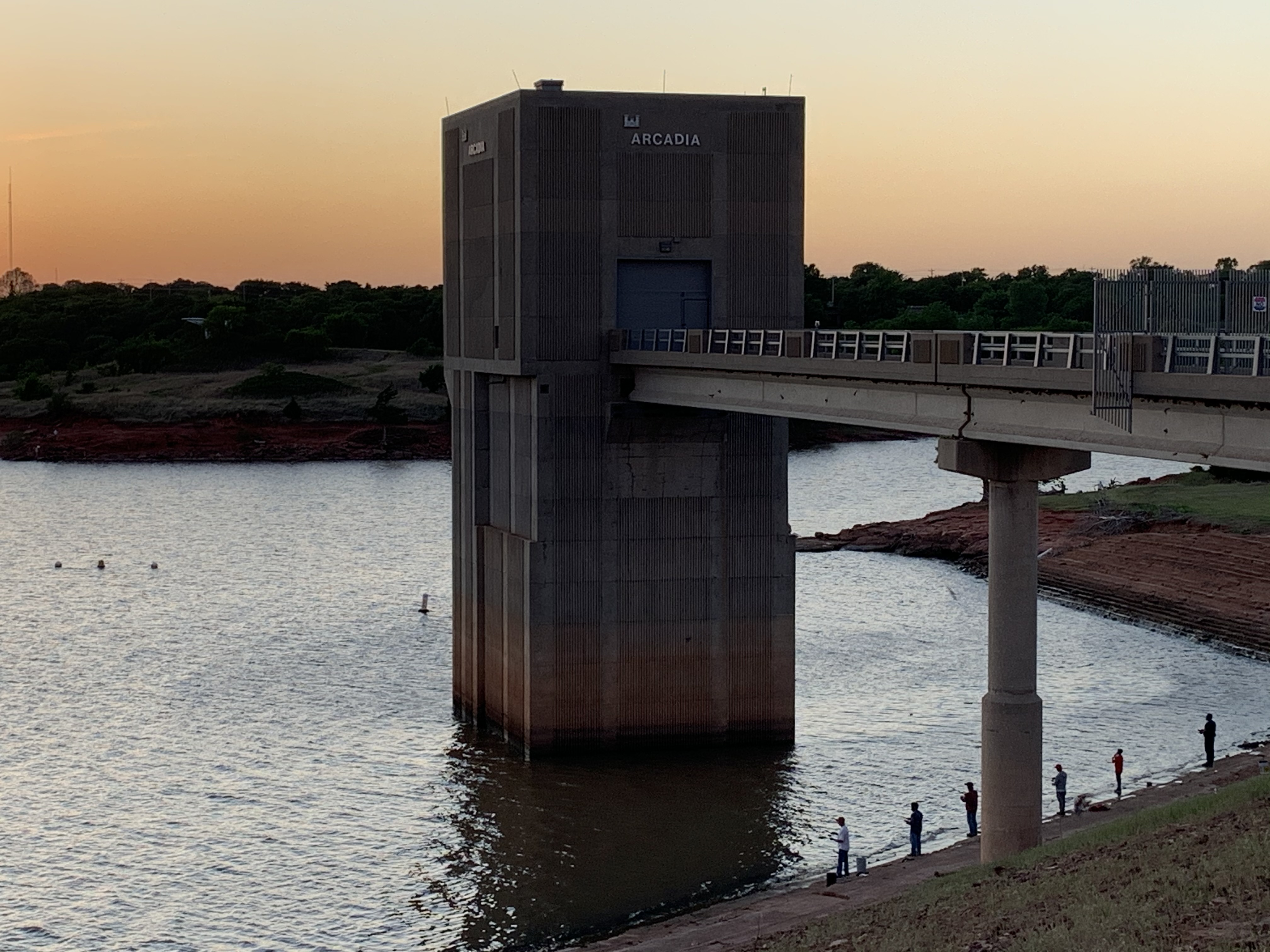 Concrete dam tower labeled 'Arcadia' beside calm lake water at sunset with people along the shoreline.