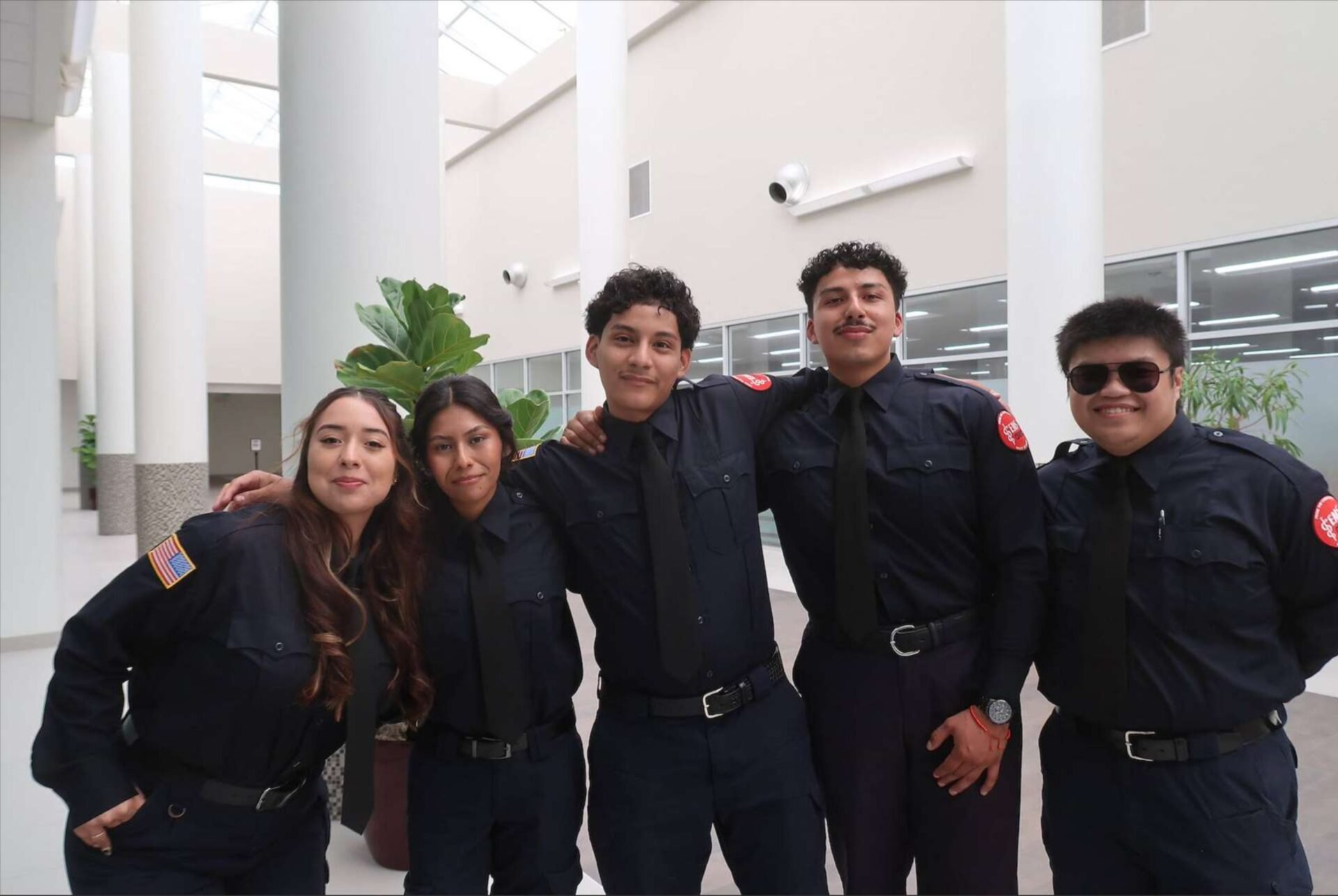 Five young adults in dark navy EMS uniforms pose together indoors, smiling.