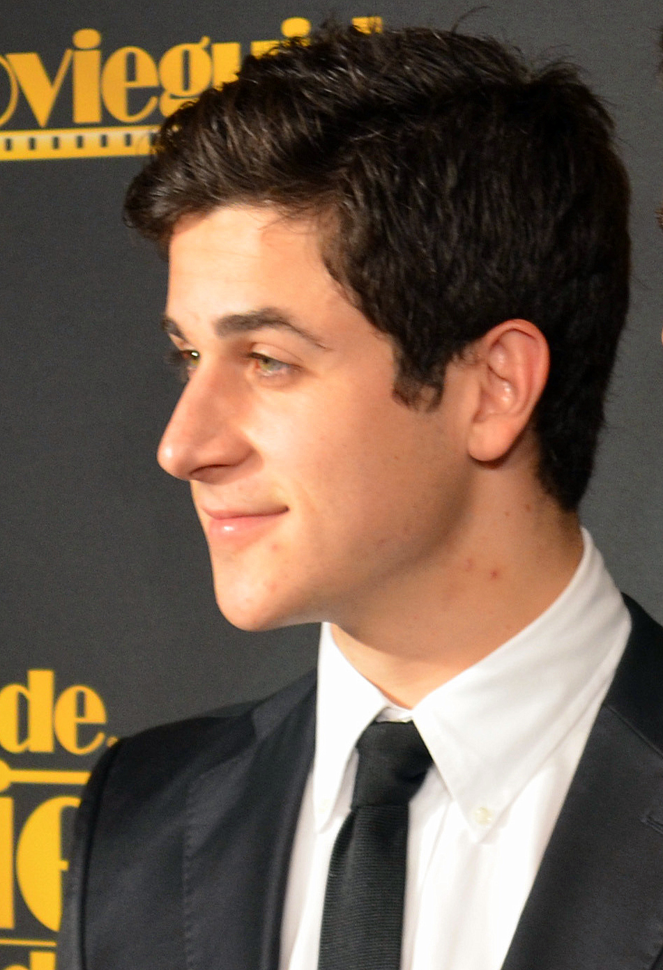 Young man in a dark suit and black tie photographed in profile at a red-carpet event.