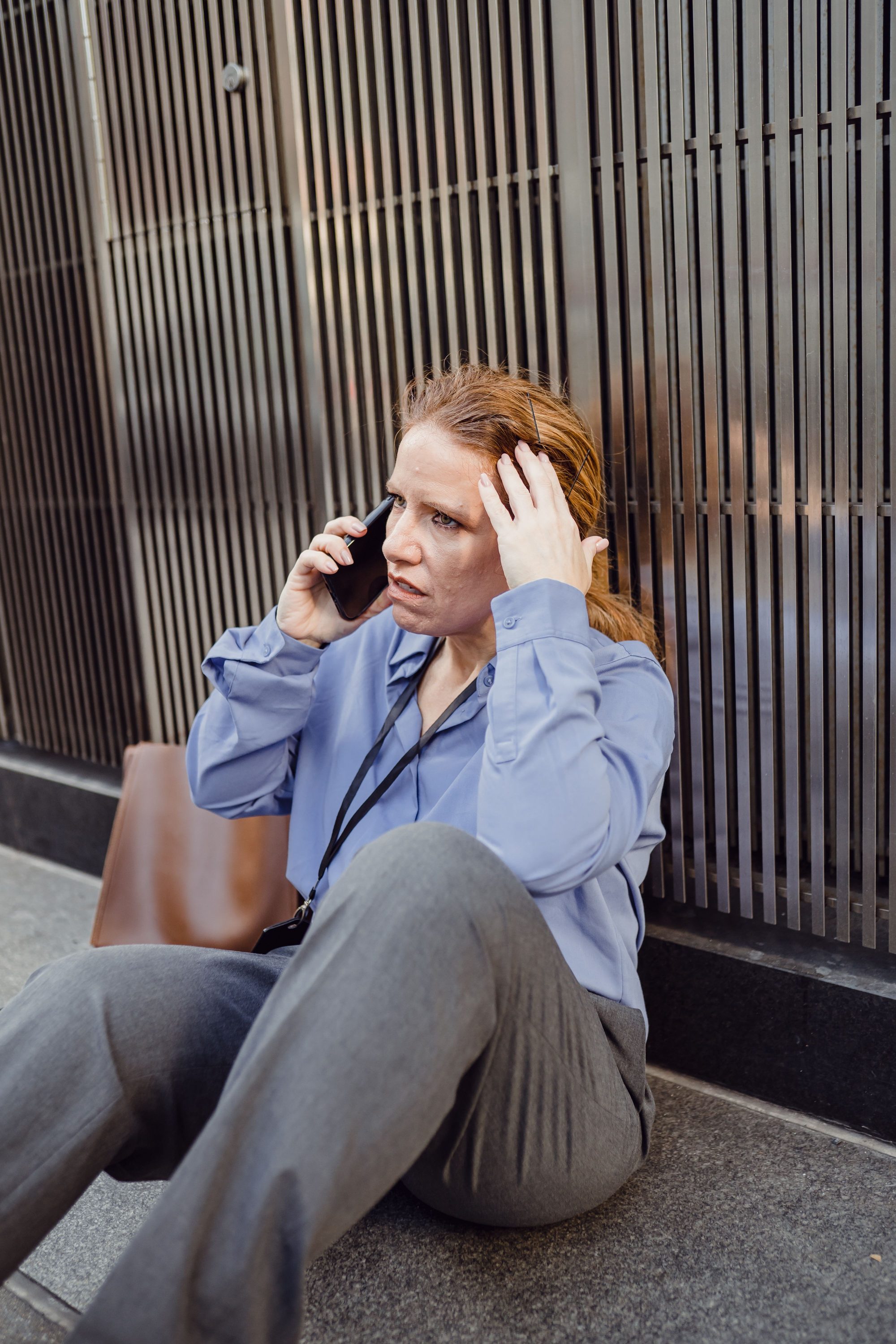 A worried woman sits on the pavement during a stressful phone call against a city backdrop.