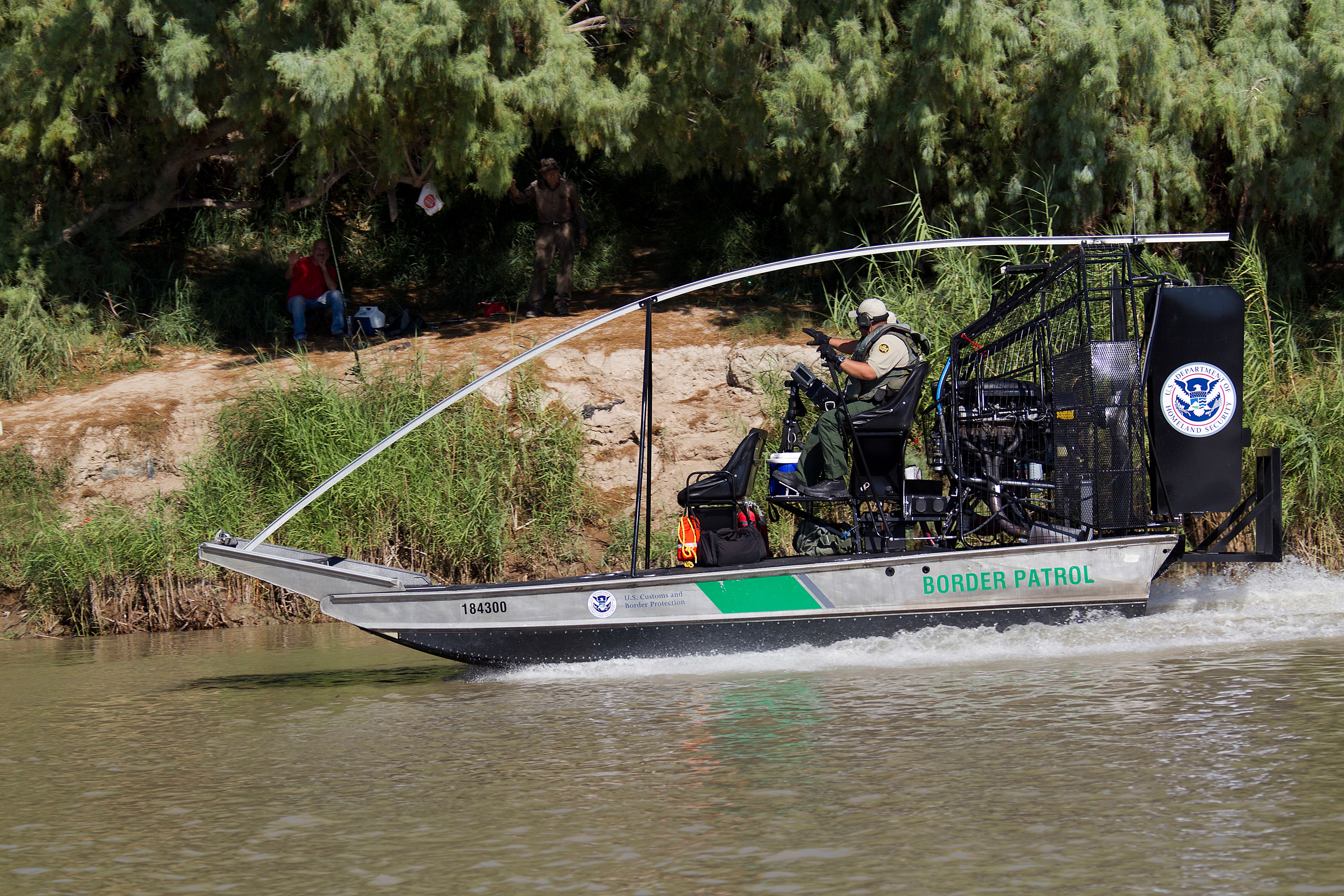 Uniformed officer at the helm of a marked U.S. Border Patrol airboat on a river.