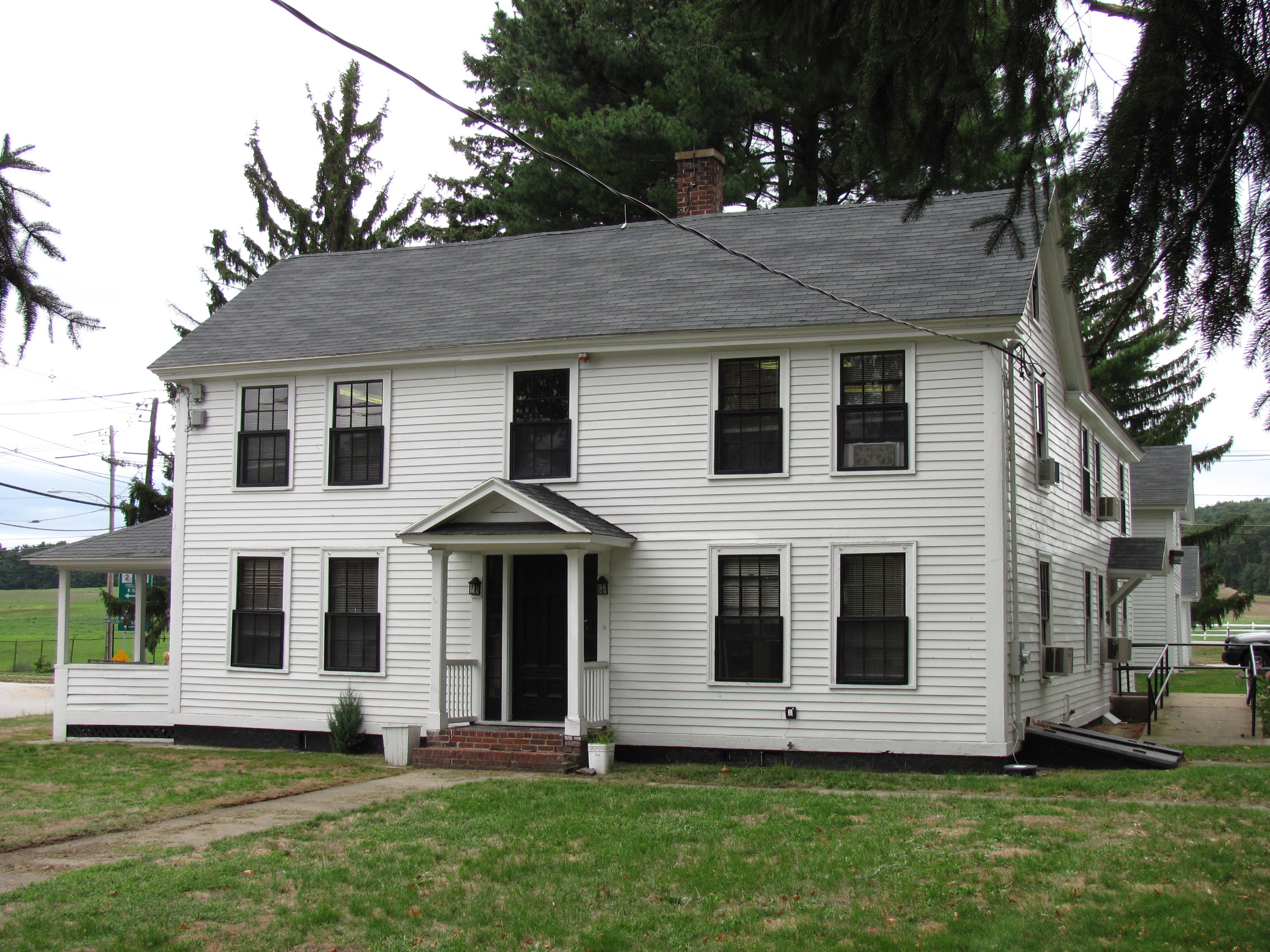 Two-story white colonial house with central portico and black-trimmed windows surrounded by trees.