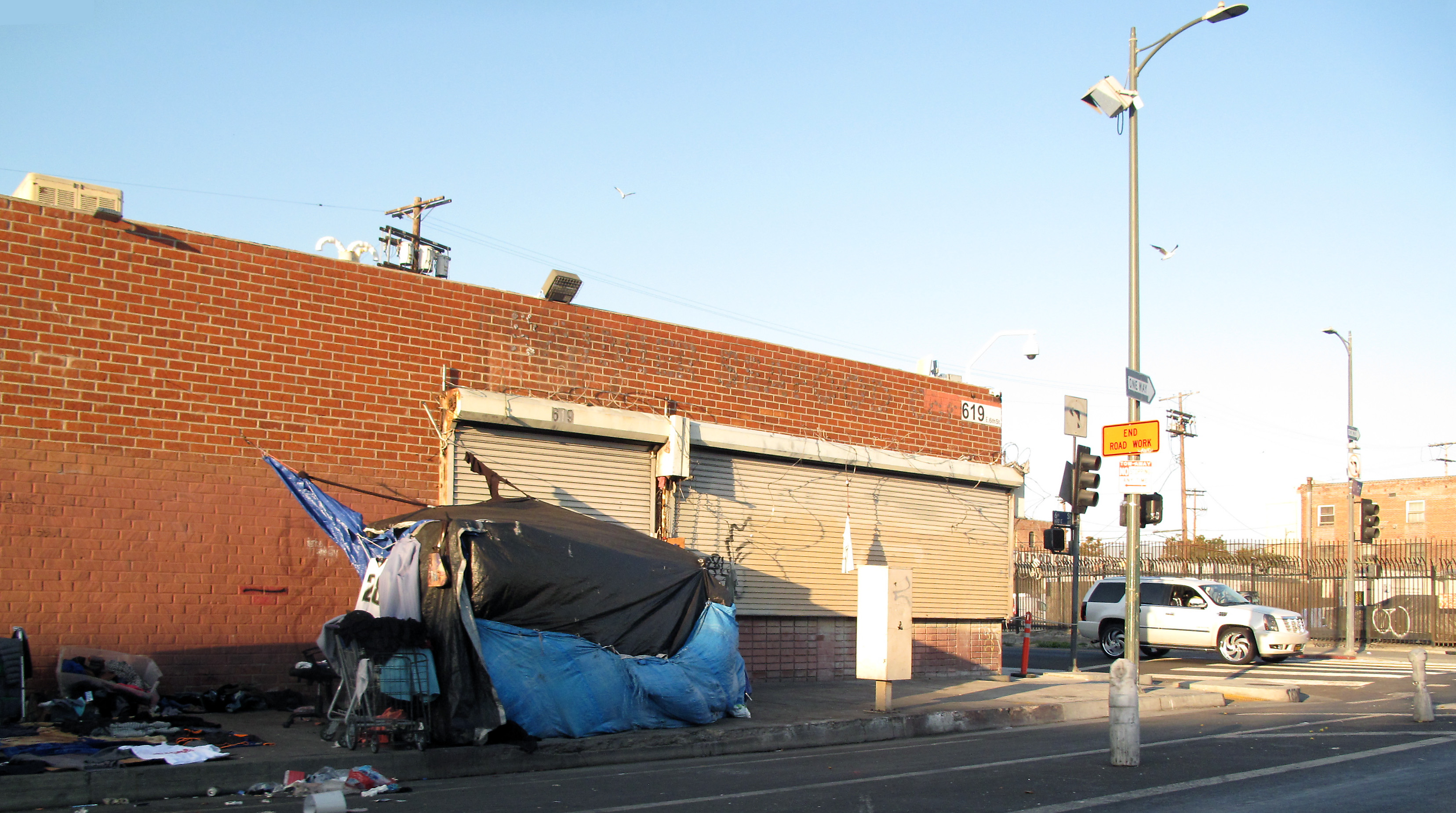 Tarp-covered encampment with scattered debris on a Skid Row sidewalk corner.
