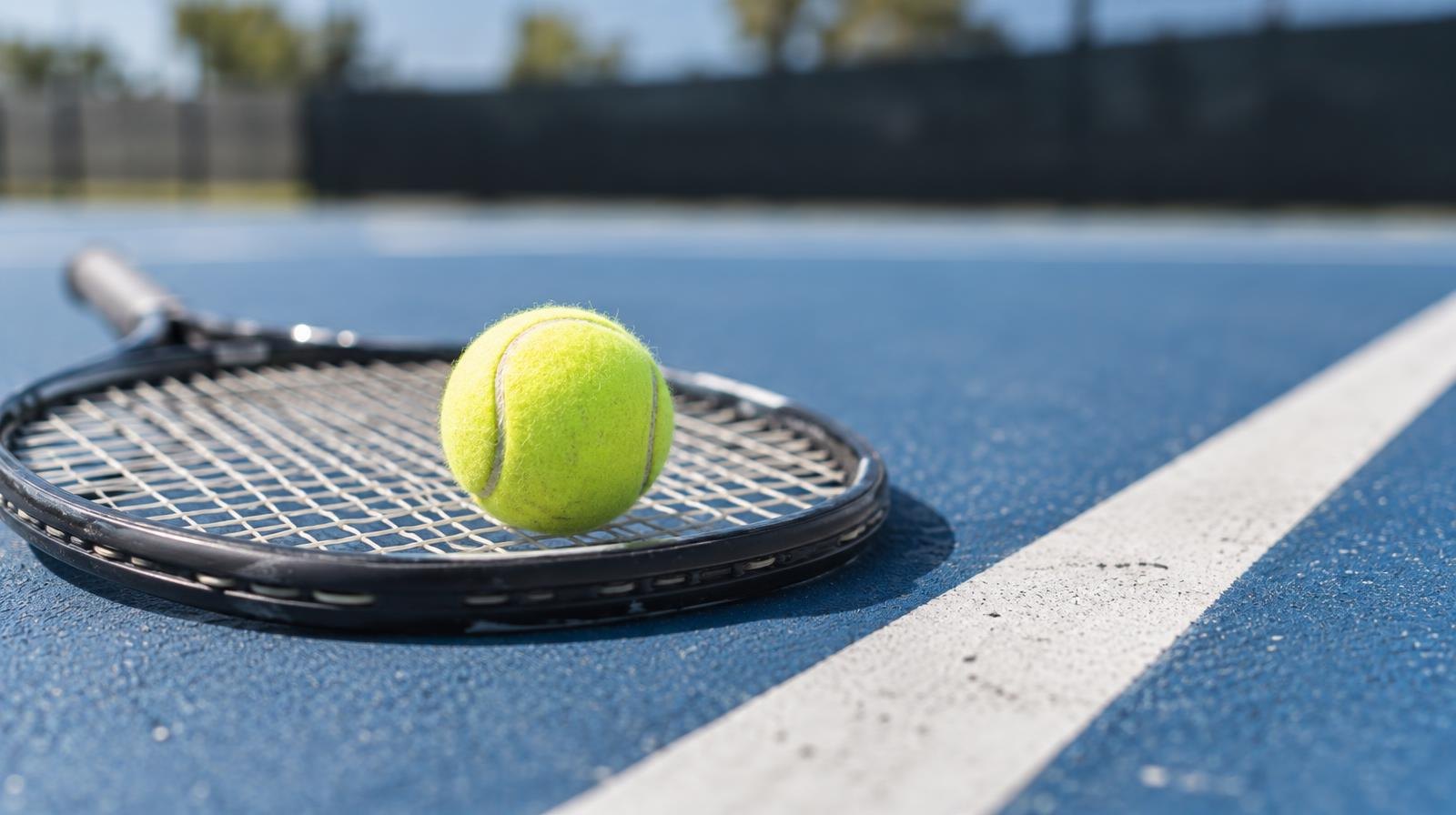 Yellow tennis ball resting on a tennis racket on a blue hard court surface.