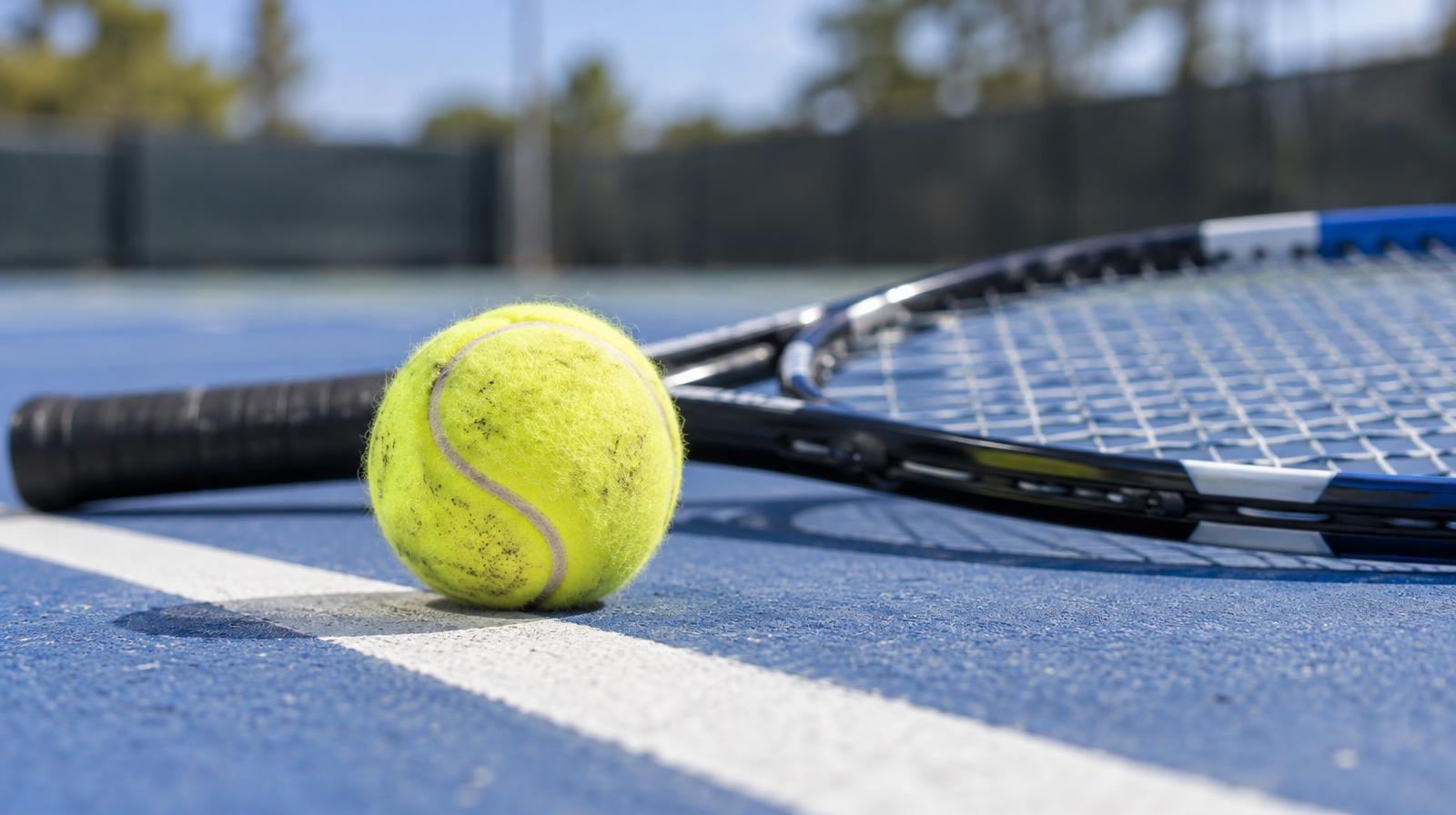 Yellow tennis ball and racket resting on a blue outdoor hard court baseline.