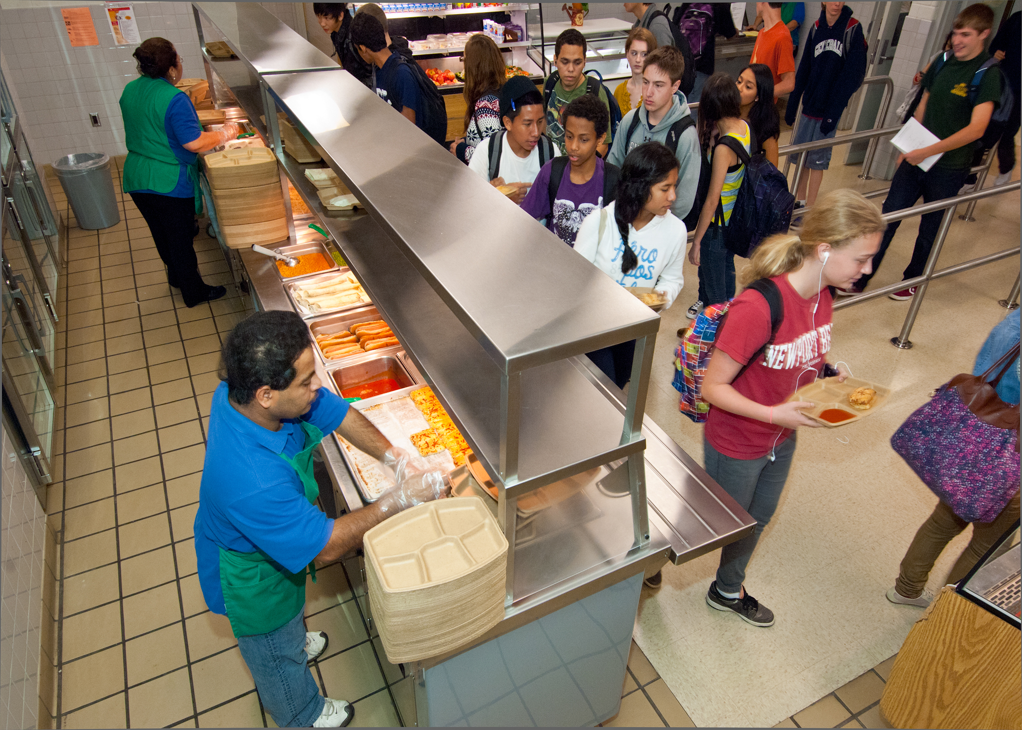 Students carry trays through a school cafeteria serving line with food workers behind the counter.