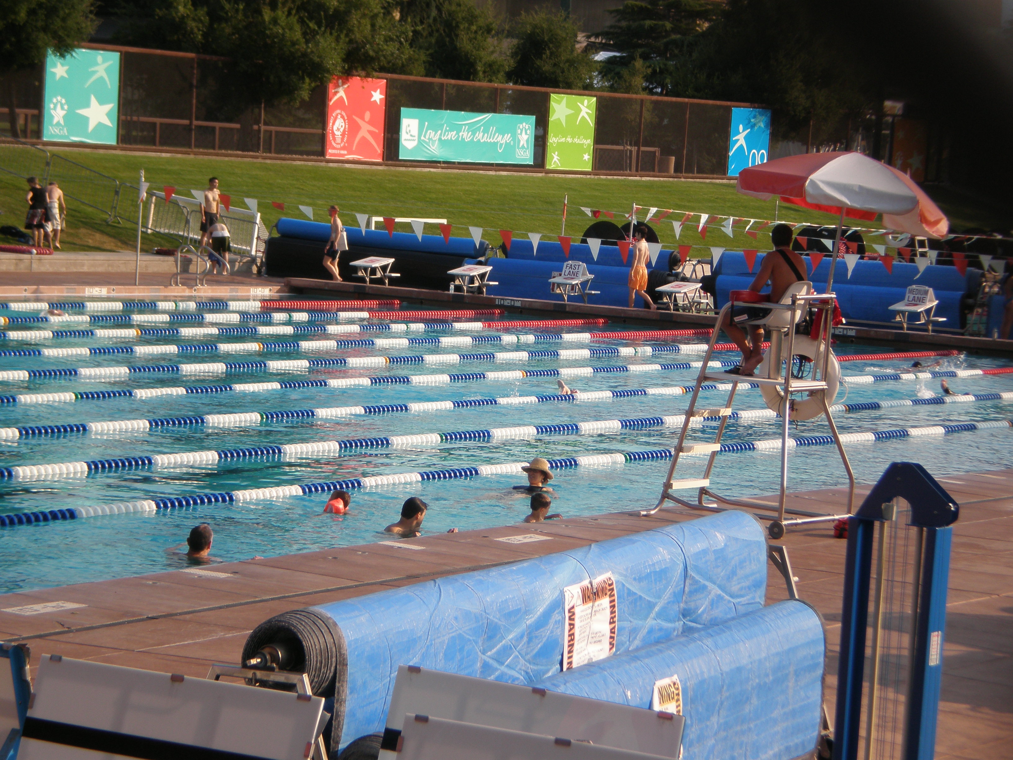 Outdoor competitive swimming pool with lane lines, swimmers, and a lifeguard stand.