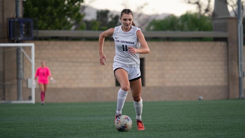 Female soccer player in white number 11 jersey dribbles a ball on turf.